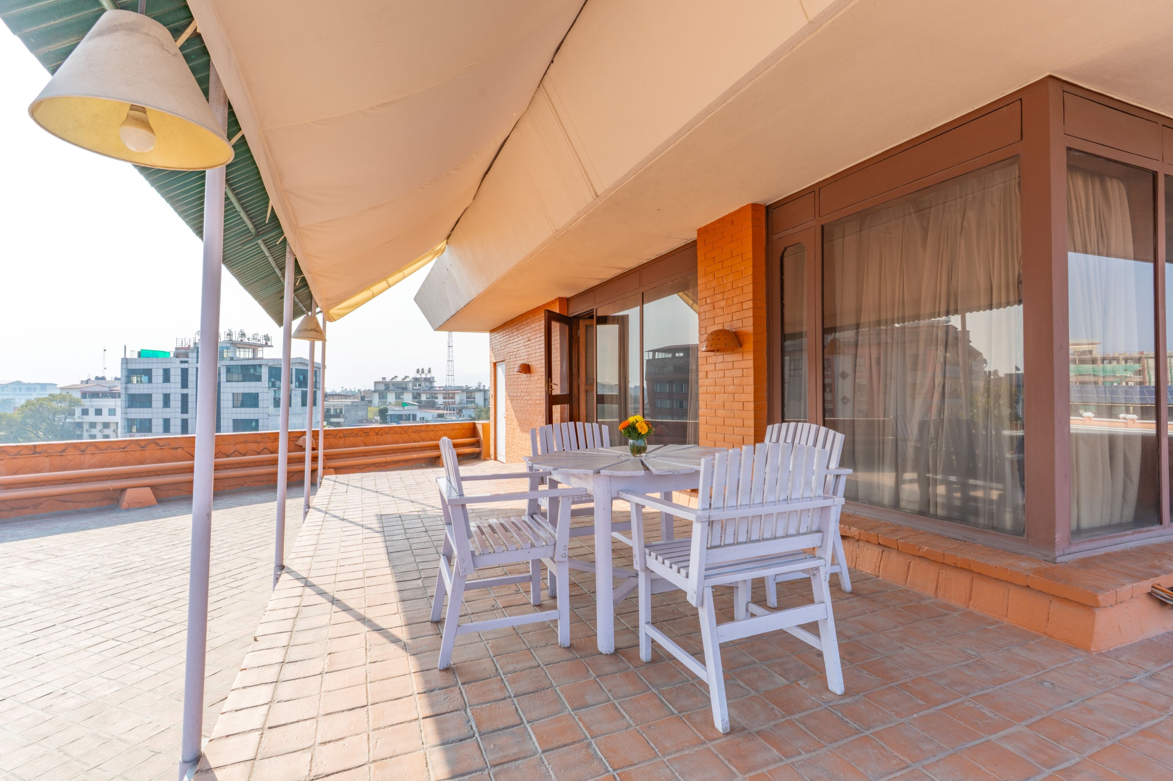 A terrace with a table and chairs, a large window with curtains, and a view of buildings in the background.