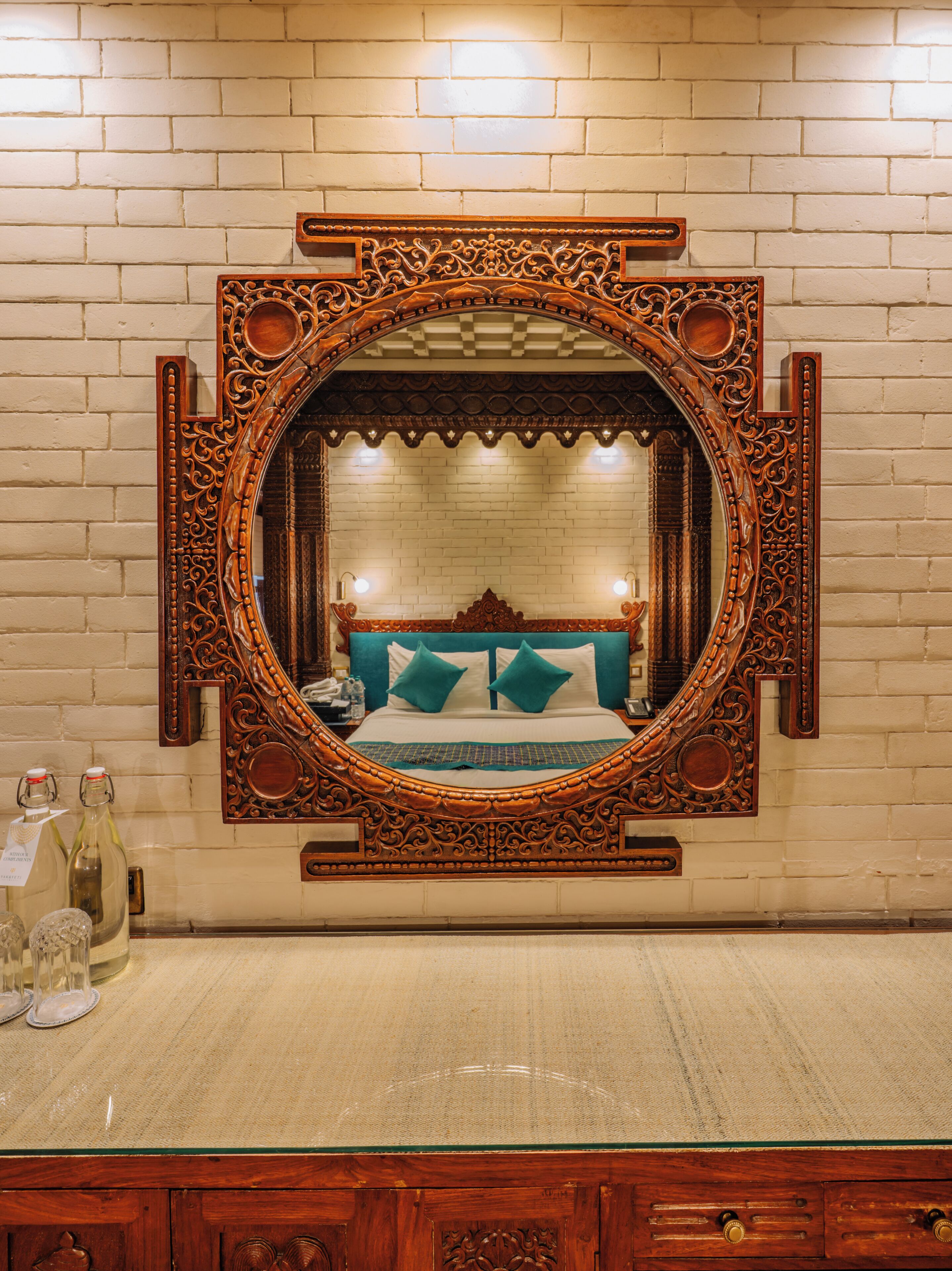 A mirror with ornate wooden carvings, a bed with teal pillows, and a brick wall.