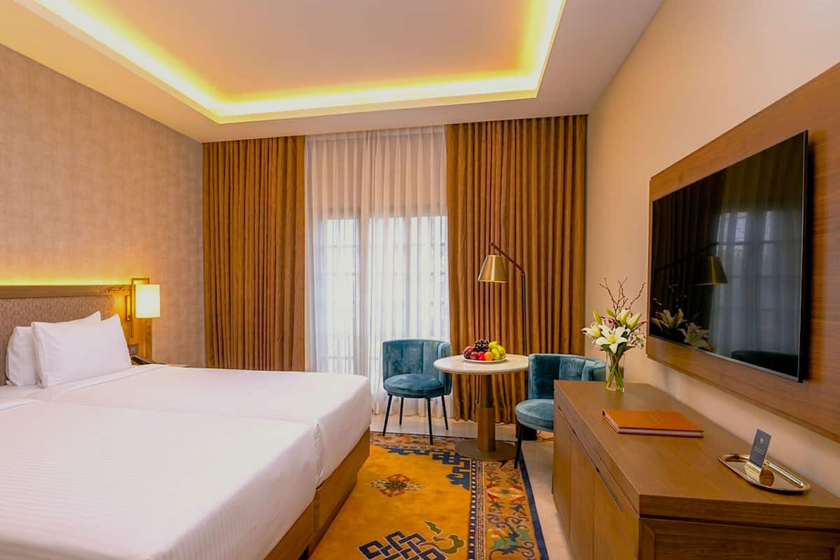 Ein Hotelzimmer mit einem großen Bett, einem Schreibtisch, einem Sessel, einem runden Tisch mit Obst und einem an der Wand befestigten Fernseher.