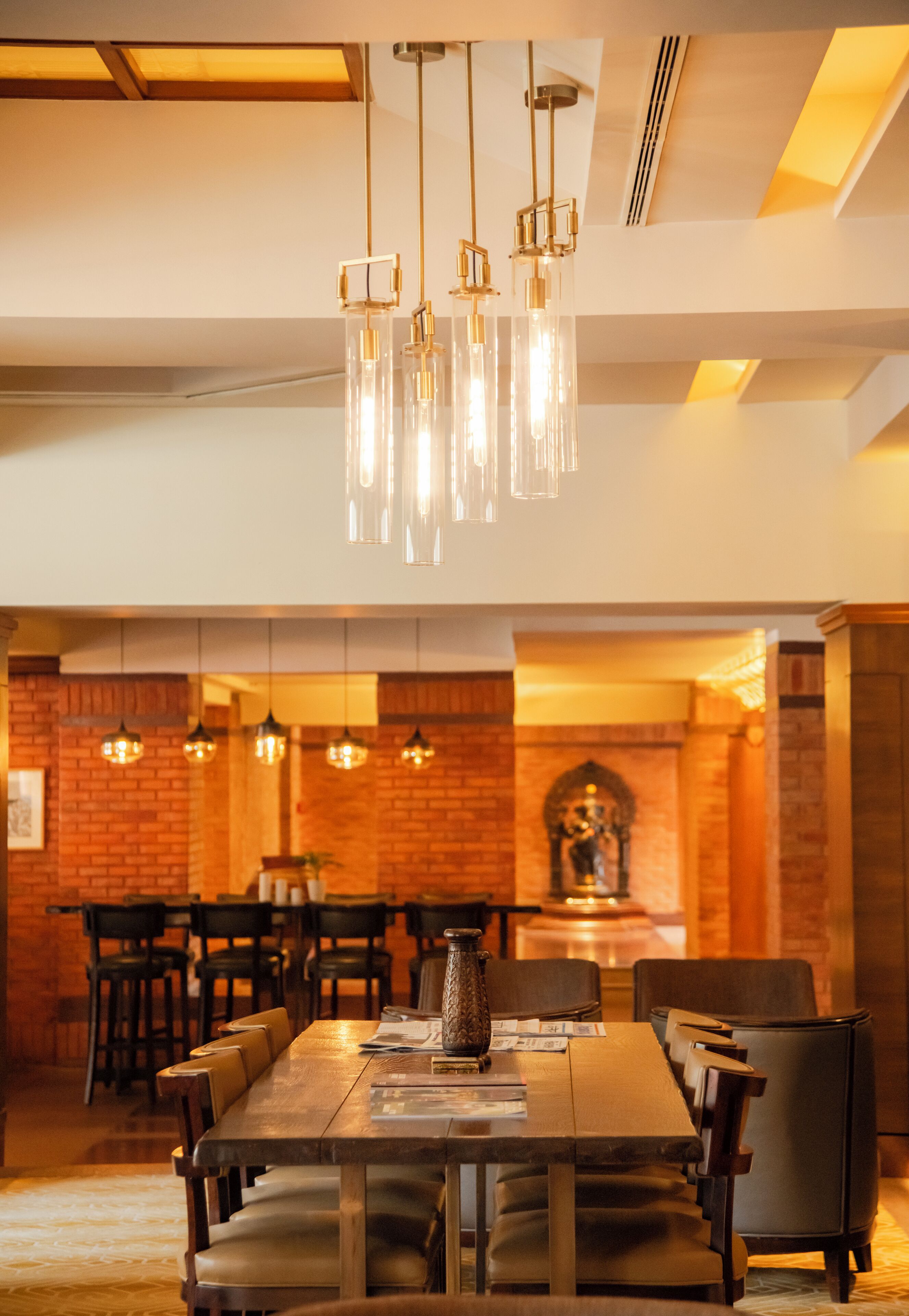 A dining area with a long wooden table, multiple chairs, hanging pendant lights, and a brick wall with a statue.