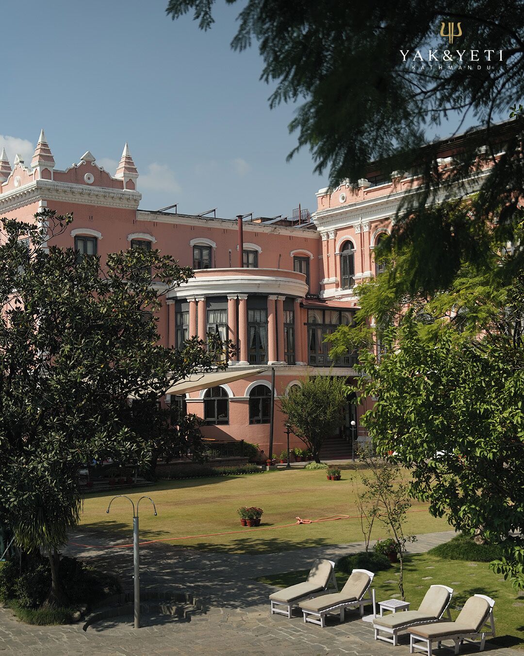 A pink building with a circular central section and a garden with lounge chairs.