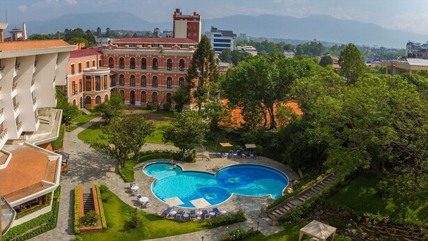 Zona de piscina con una piscina en forma de corazón, rodeada de sillones de descanso y sombrillas, con un edificio grande al fondo.