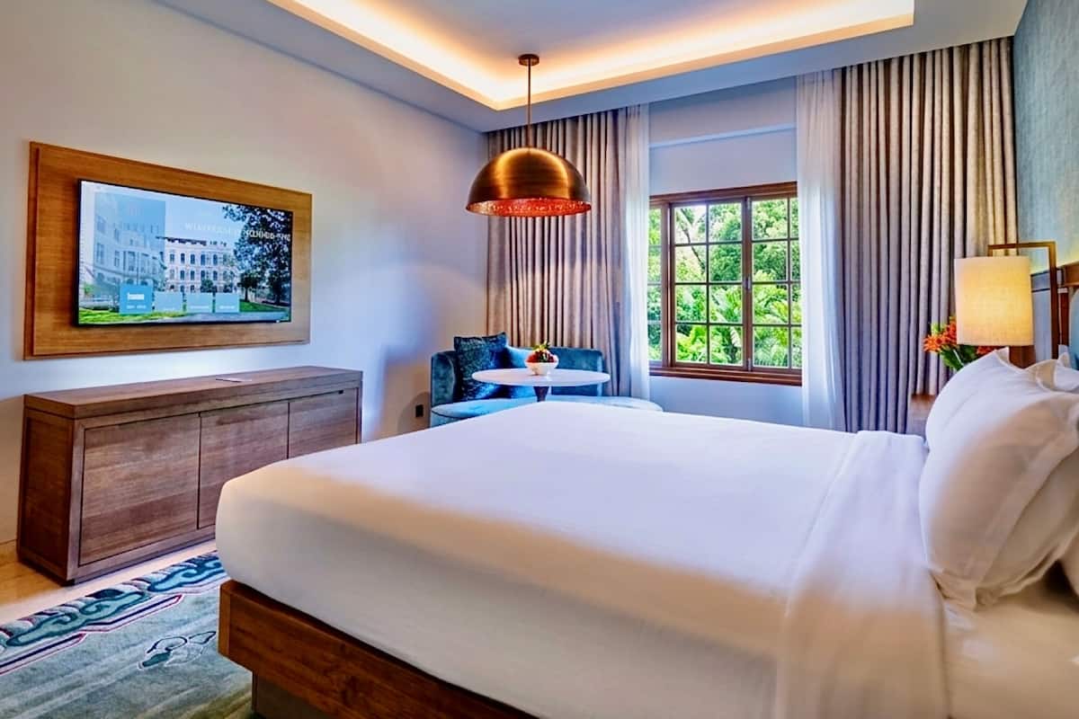 Ein Hotelzimmer mit einem großen Bett, einer Holzkommode, einem an der Wand montierten Fernseher, einem runden Tisch mit einer Vase und einem Fenster mit Vorhängen.