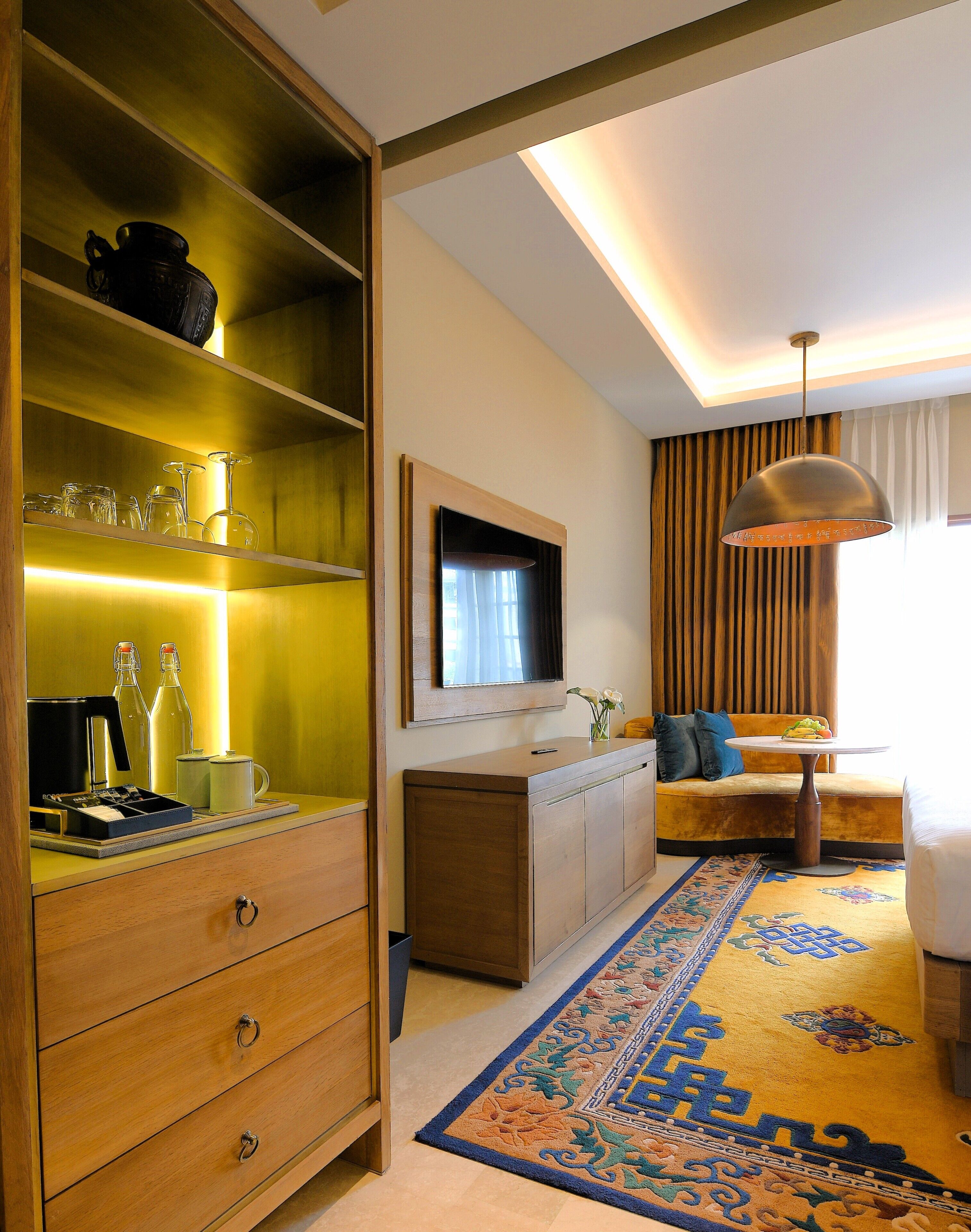 A hotel room with a wooden cabinet, a television mounted on the wall, a sofa, a small table, and a patterned rug.