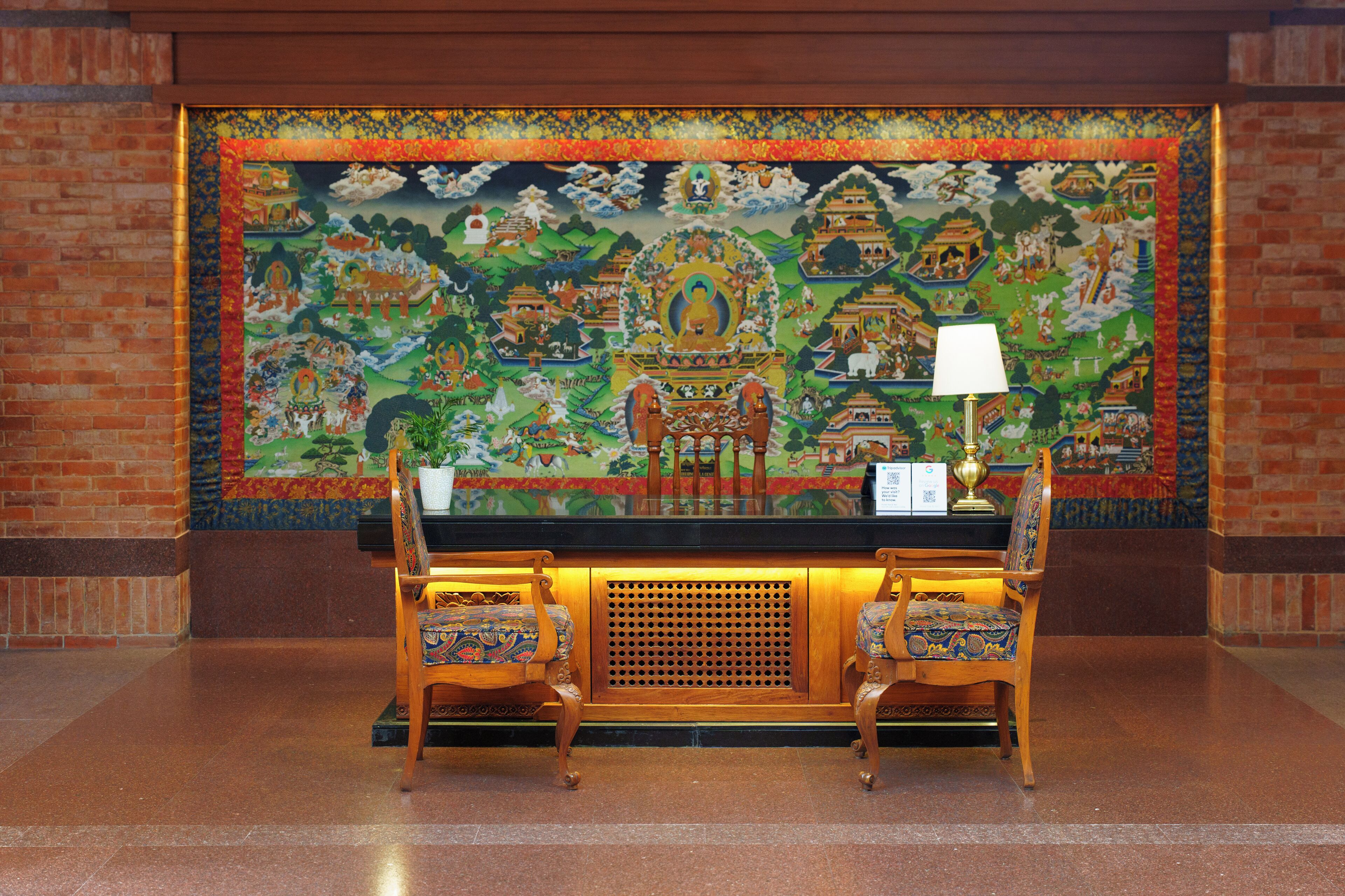 A reception desk with two chairs, a lamp, and a large tapestry depicting traditional architecture and figures.