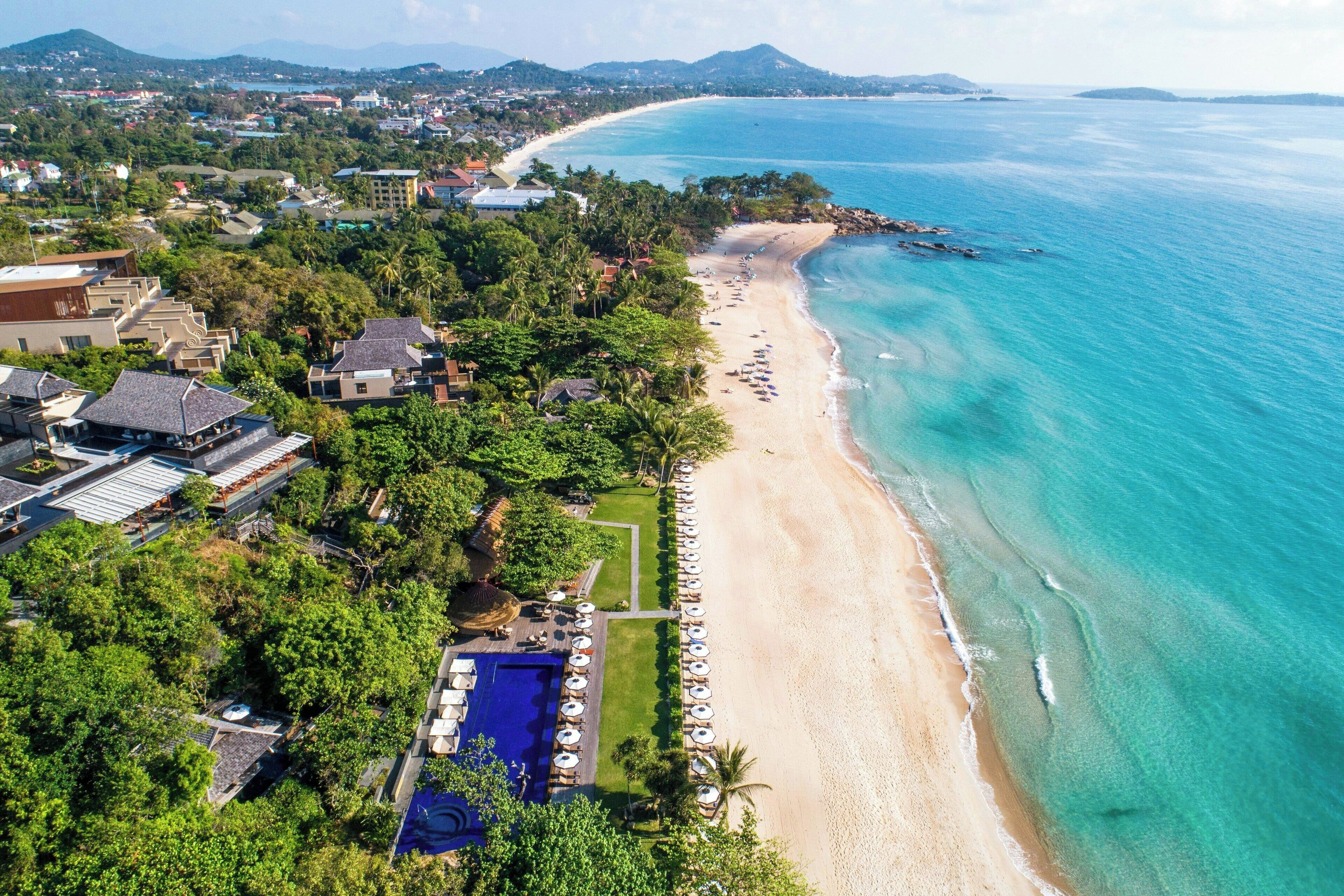 An aerial view of a coastal resort with a beach, a swimming pool, and surrounding greenery.