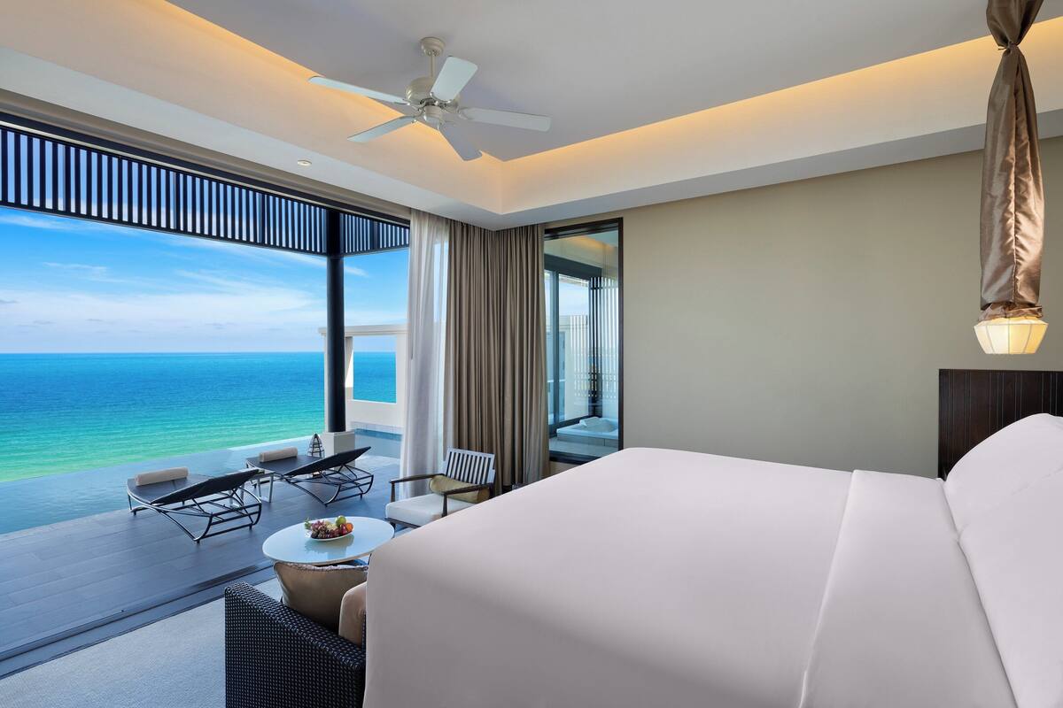 A hotel room with a large bed, a ceiling fan, a dining area with a table and chairs, and a view of the ocean.