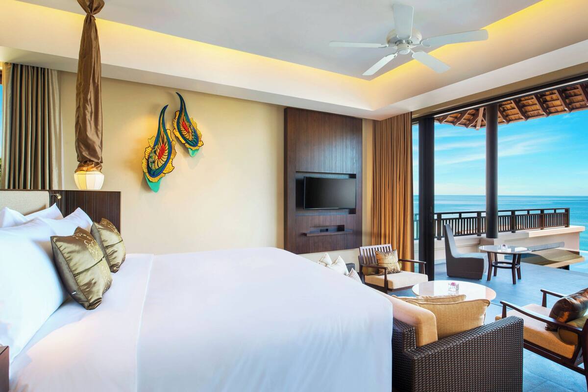 A hotel room with a large bed, a TV, a balcony with seating, and a view of the ocean.
