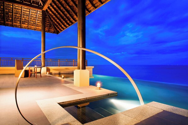 A serene pool area with a clear view of the ocean at dusk, a wooden deck, and a seating area with a table and chairs.
