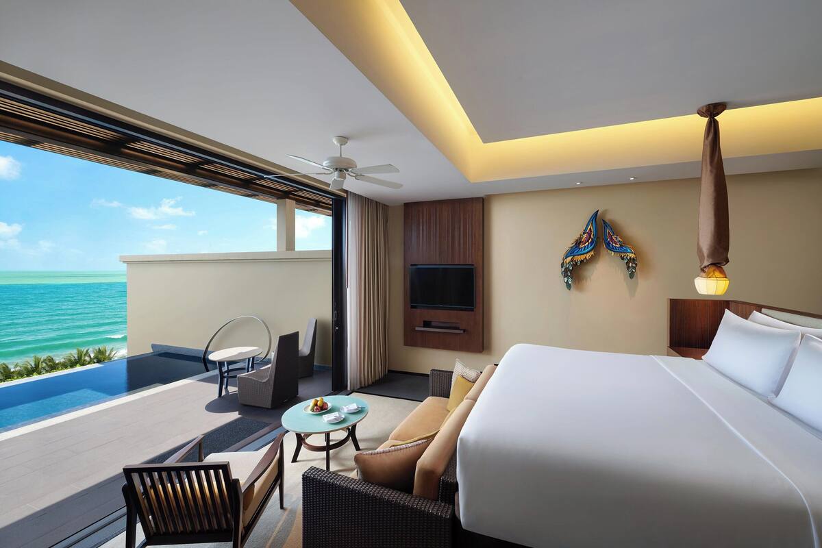 A hotel room with a bed, a sofa, a small table, a chair, a TV, and a balcony with a view of the ocean.