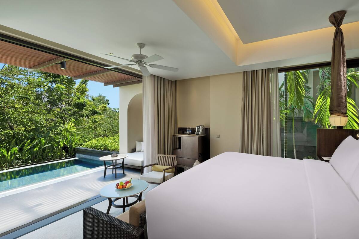 A hotel room with a bed, a small table with fruit, a chair, a desk, a ceiling fan, a minibar, and a view of a swimming pool and greenery.