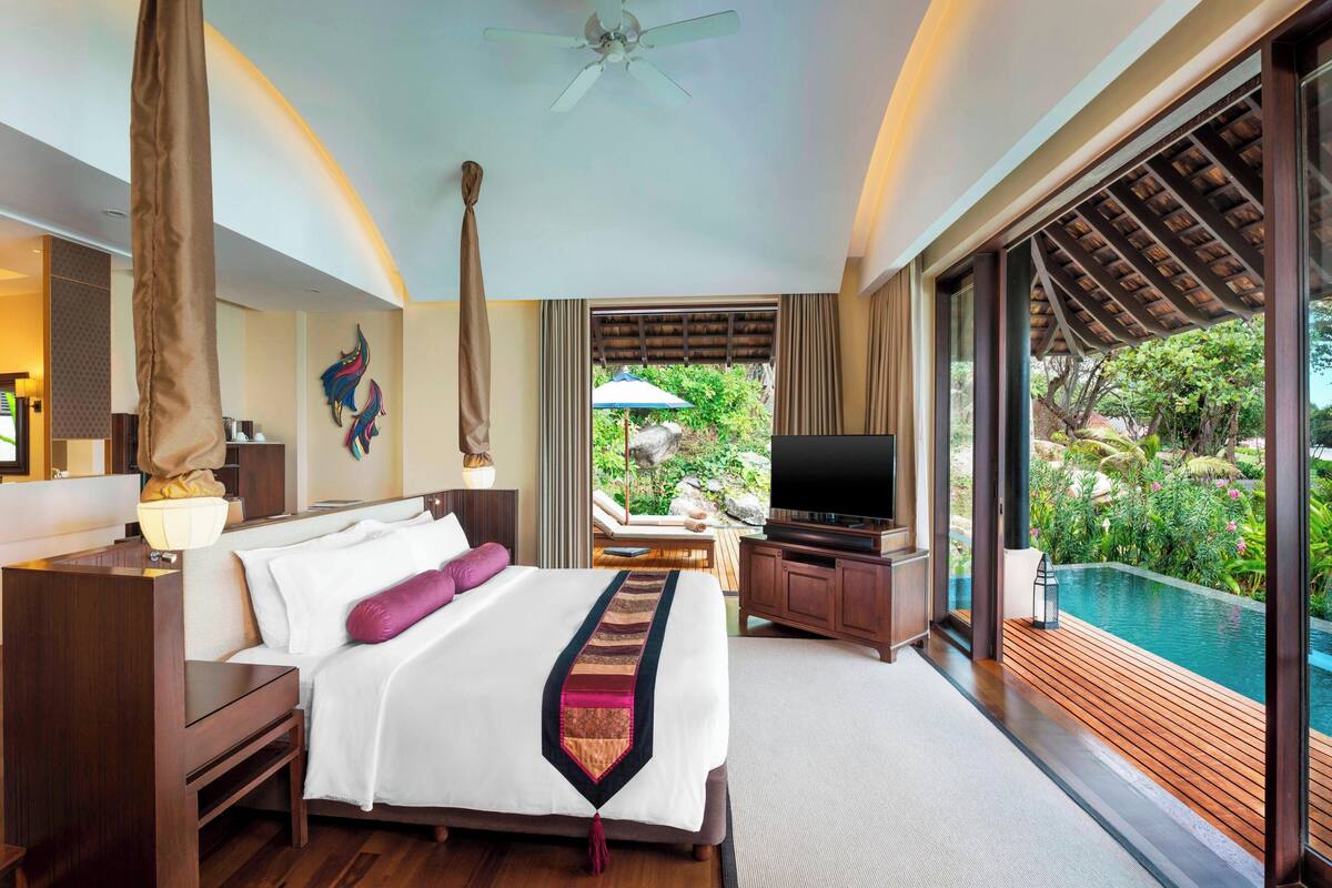 A hotel room with a large bed, a television, a wooden side table, and a view of a swimming pool.