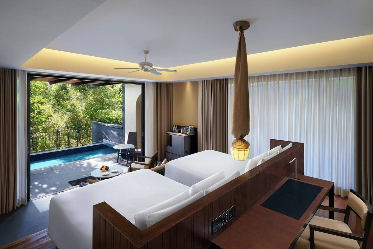A hotel room with a large bed, a desk, a chair, a small table, a ceiling fan, and a view of a swimming pool and greenery.