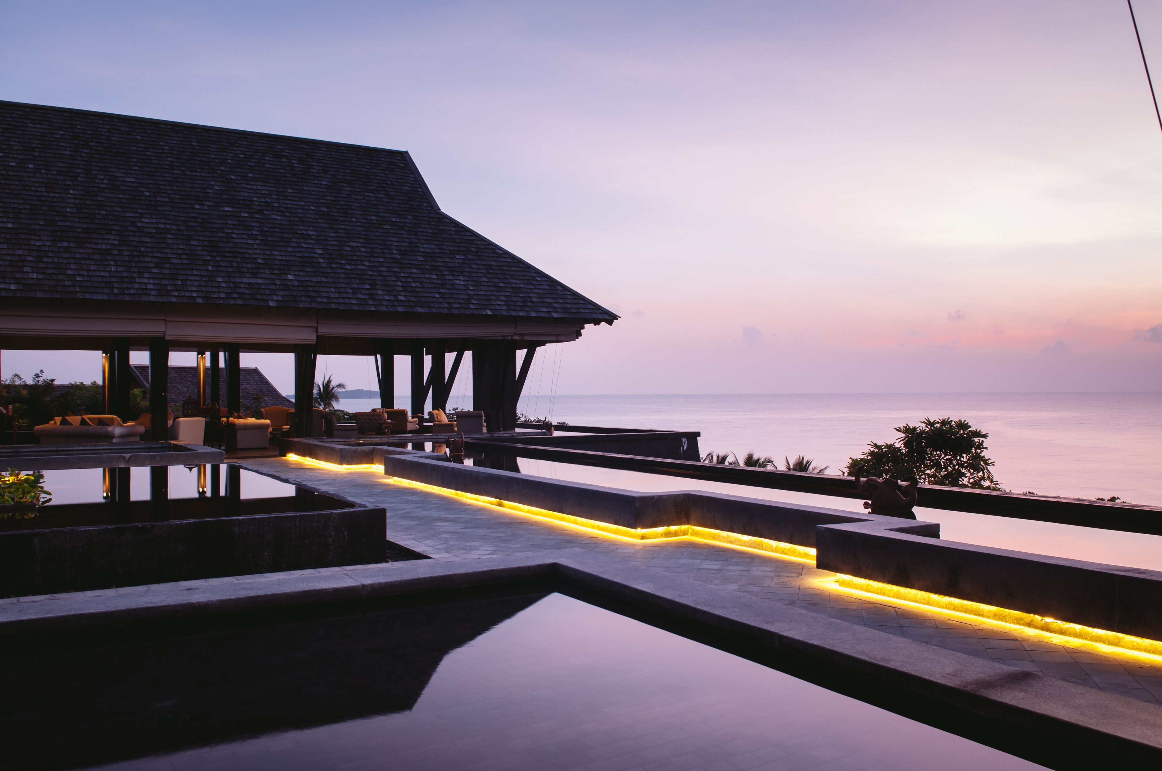 A modern outdoor lounge area with a pool, seating, and a view of the sea at dusk.
