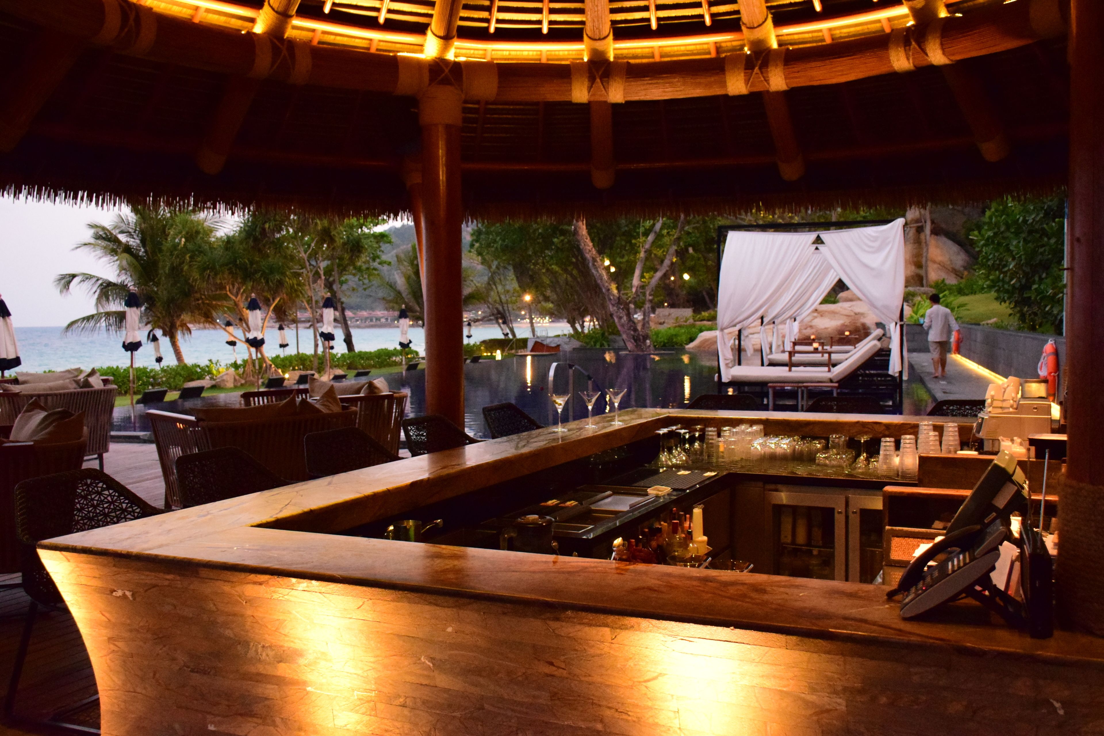 Zona de bar al aire libre con piscina, mobiliario de madera y vista al océano.