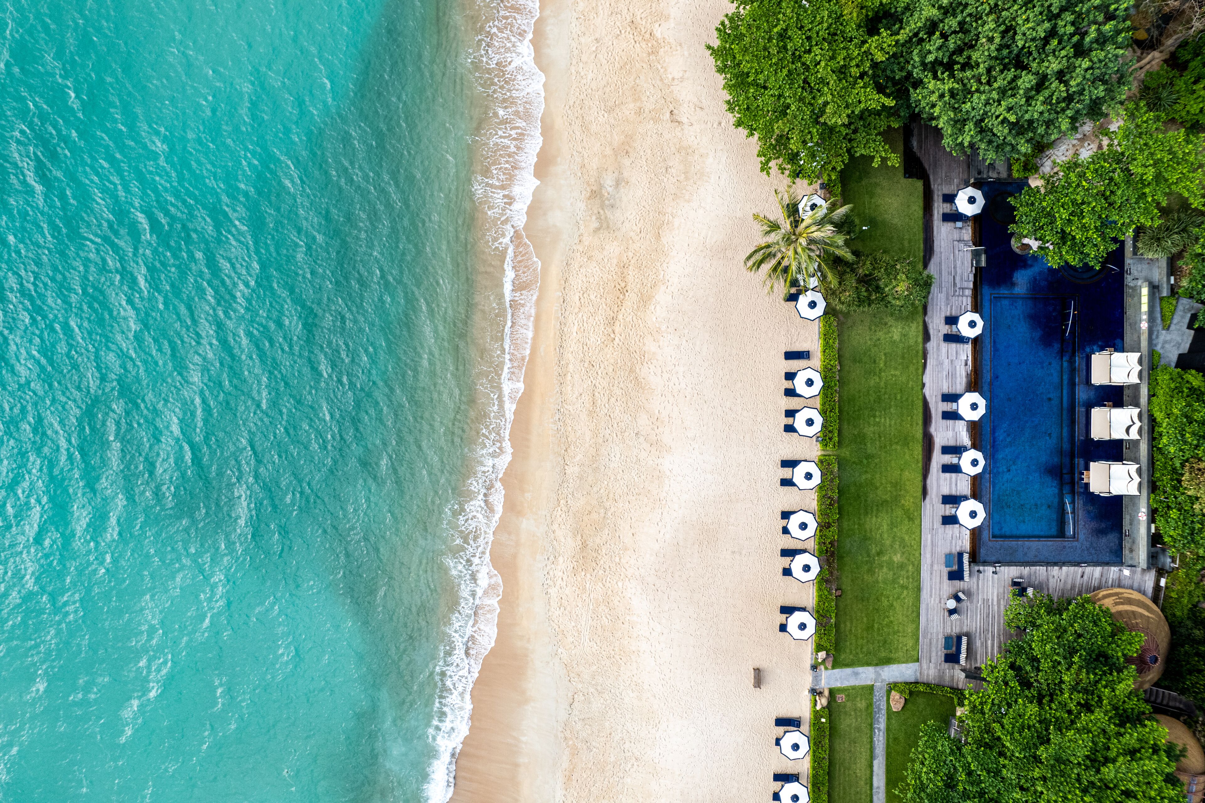 An aerial view of a beachside resort with a swimming pool, lounge chairs, and a well-maintained lawn.