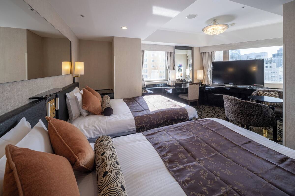 A hotel room with two beds, a desk, a television, and a large window with a city view.