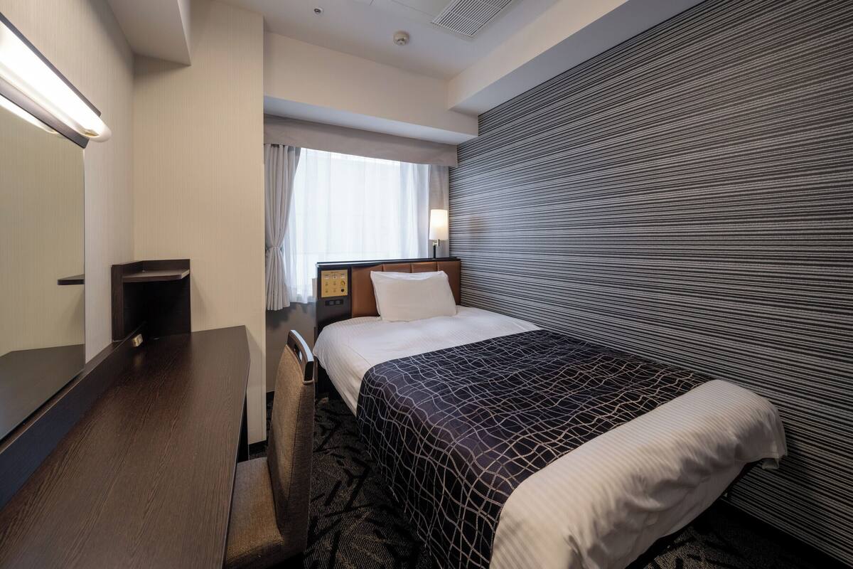 Ein Hotelzimmer mit Bett, Schreibtisch, Stuhl, Nachttisch und einem Fenster mit Vorhängen.