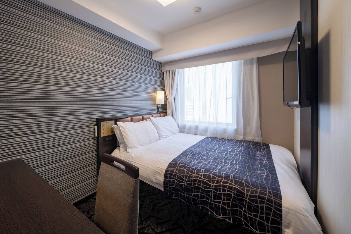 Ein Hotelzimmer mit Bett, Schreibtisch, Sessel, Fernseher und einem Fenster mit Vorhängen.