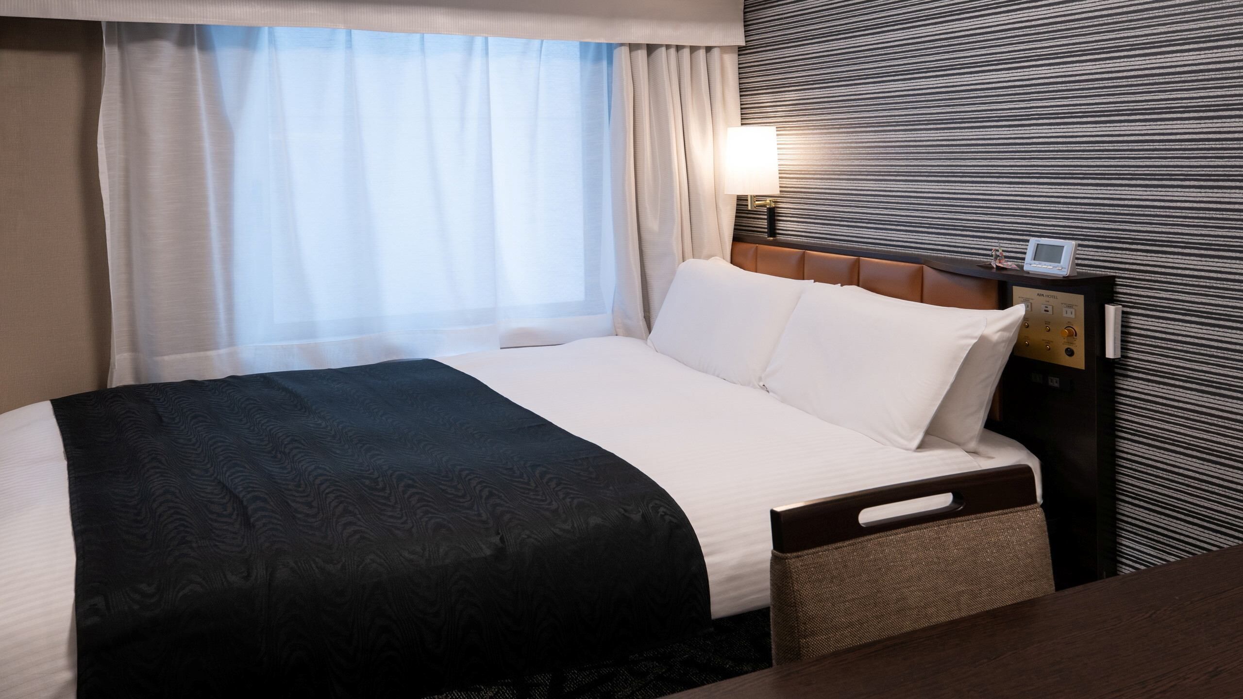 A hotel room with a bed, a bedside table, a lamp, and a control panel.