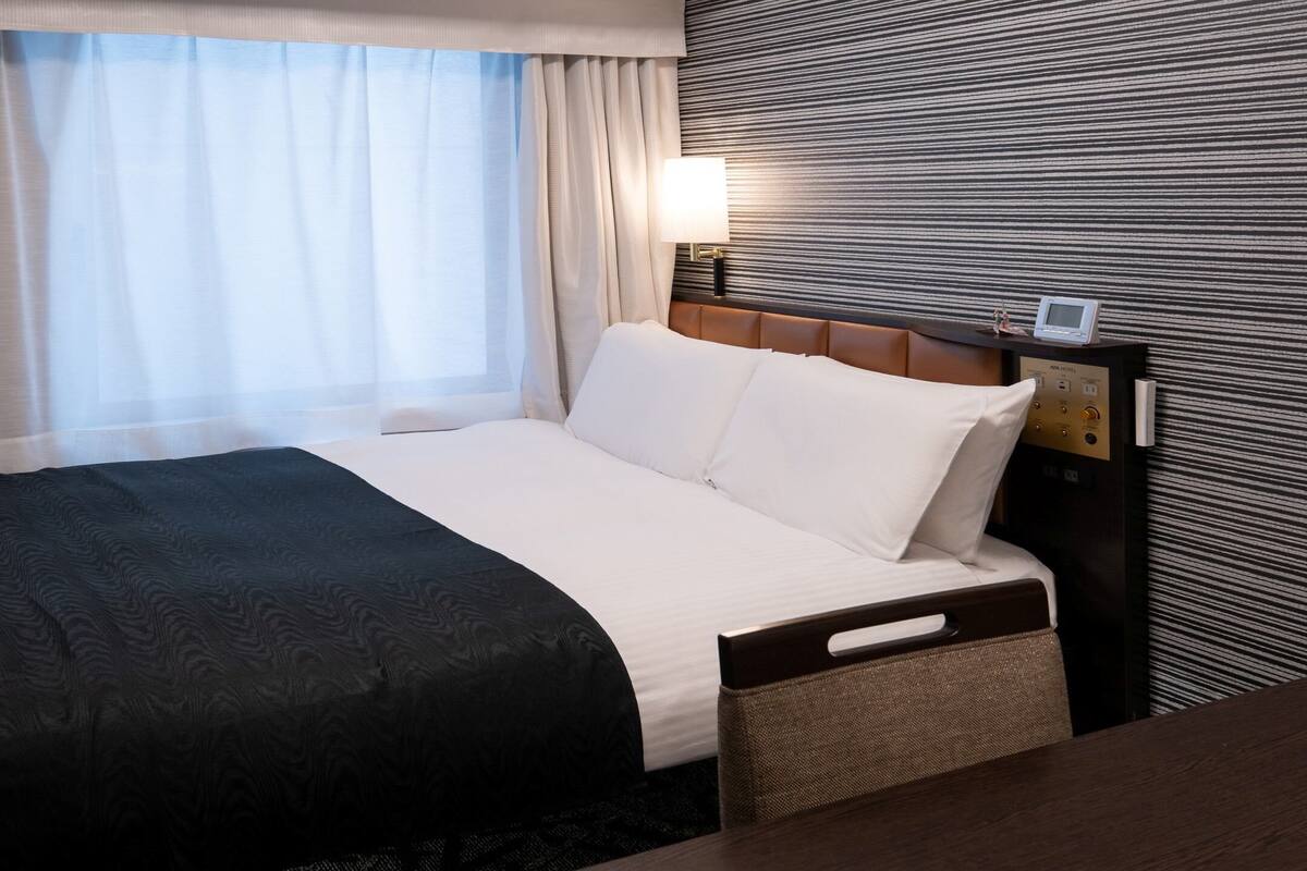 A hotel room with a bed, a bedside table, a lamp, and a control panel.