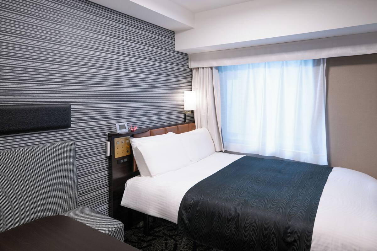 A hotel room with a large bed, a headboard with a clock, a bedside table, and a window with curtains.