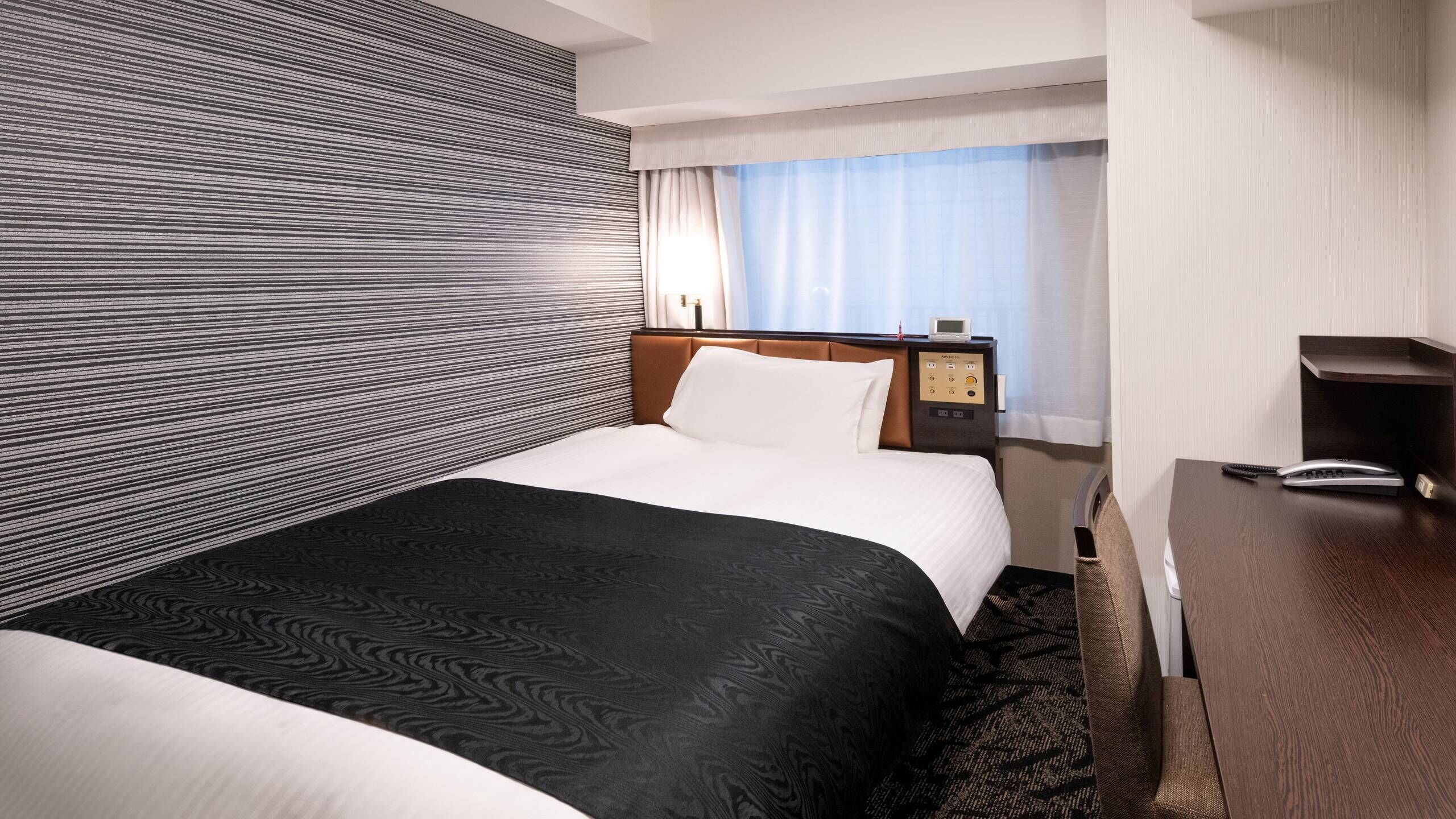 A hotel room with a bed, a bedside table with a phone, a control panel, and a window with curtains.