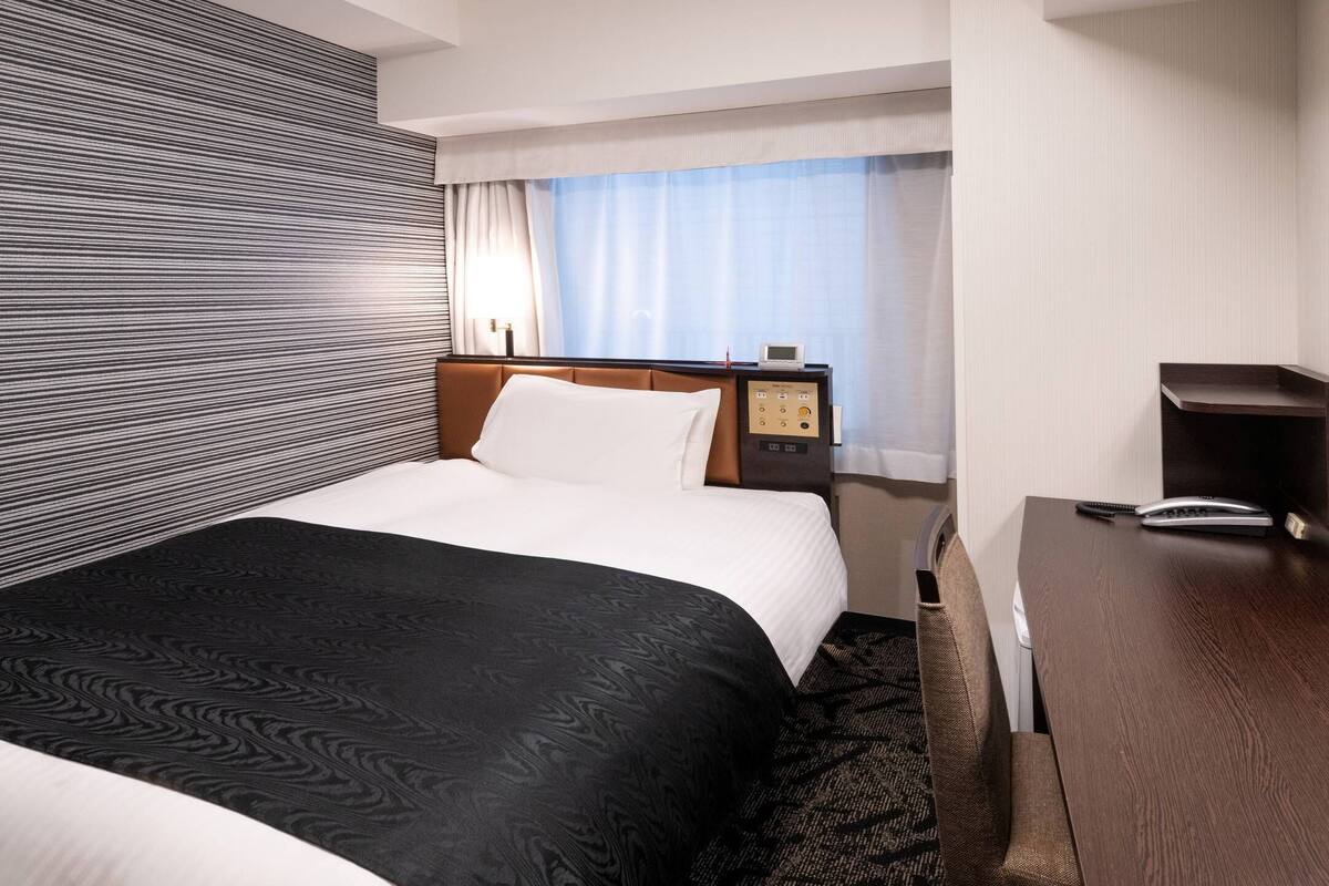 A hotel room with a bed, a bedside table with a phone, a control panel, and a window with curtains.