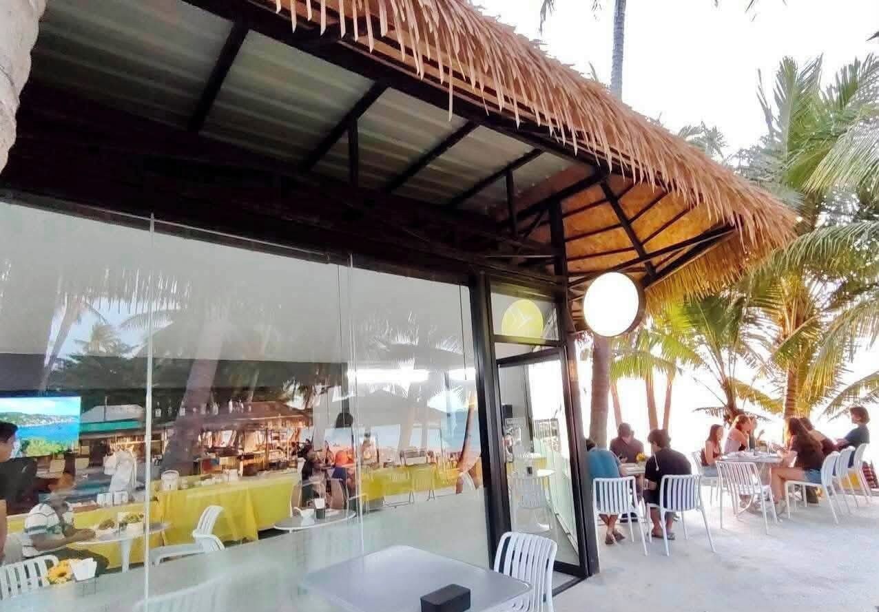 Een terras met tafels en stoelen, een rieten dak en palmbomen op de achtergrond.