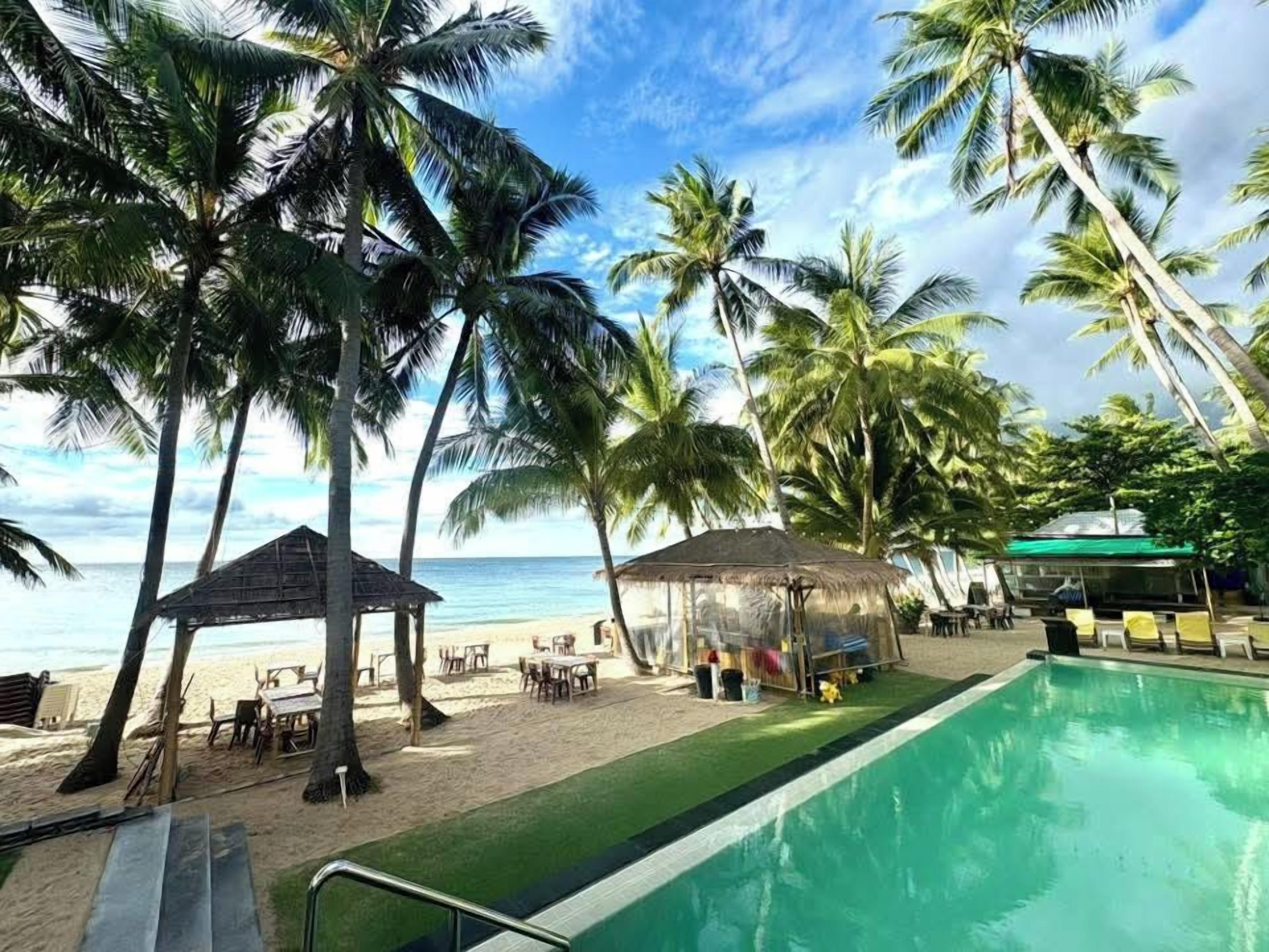 Un área de piscina con vista a la playa, palmeras y un pabellón.