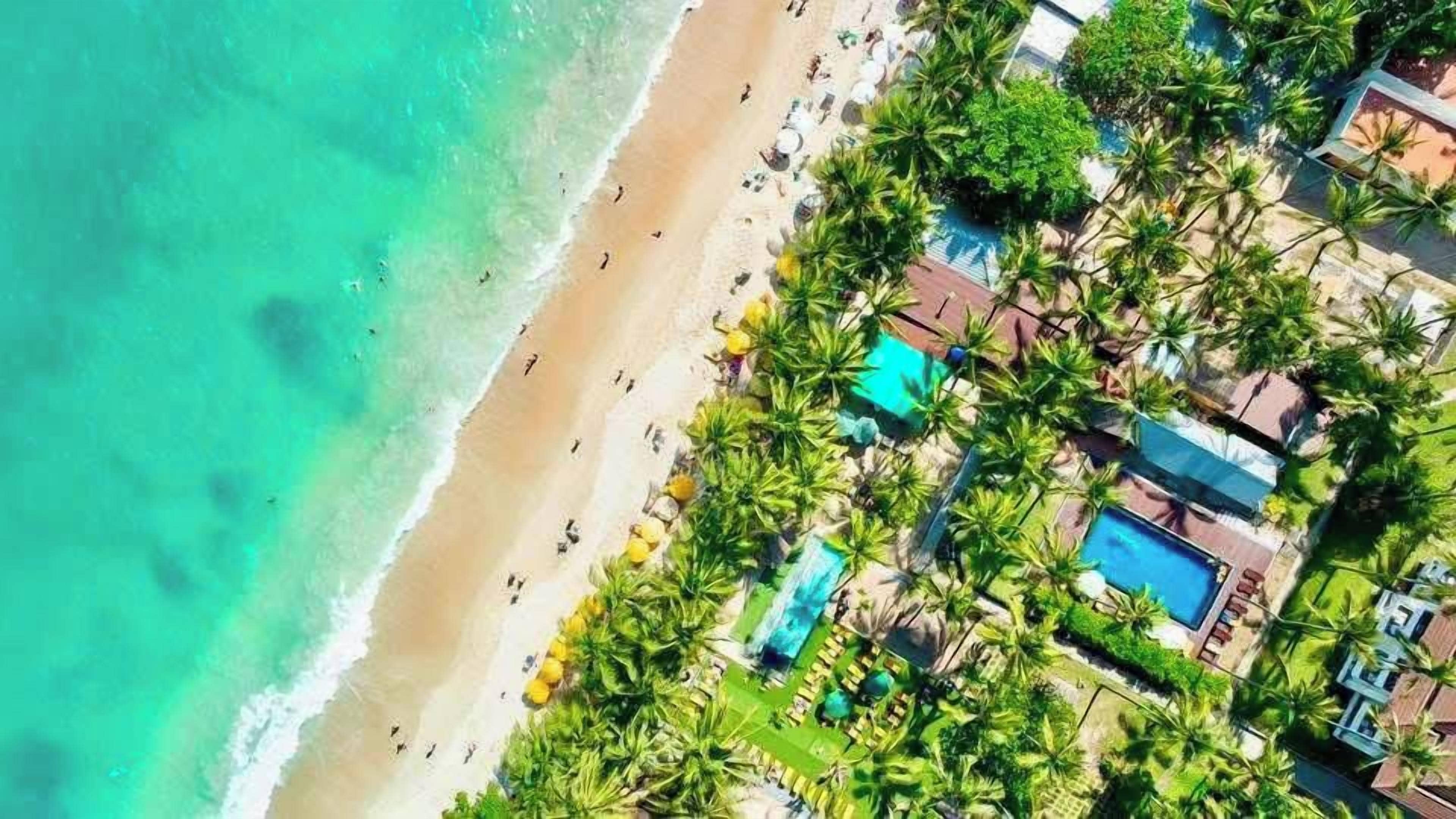 Vista aérea de um resort na praia, com piscina, palmeiras e pessoas na praia.