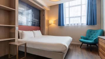 A hotel room with a bed, a desk, a chair, a wall-mounted lamp, and a window with curtains.