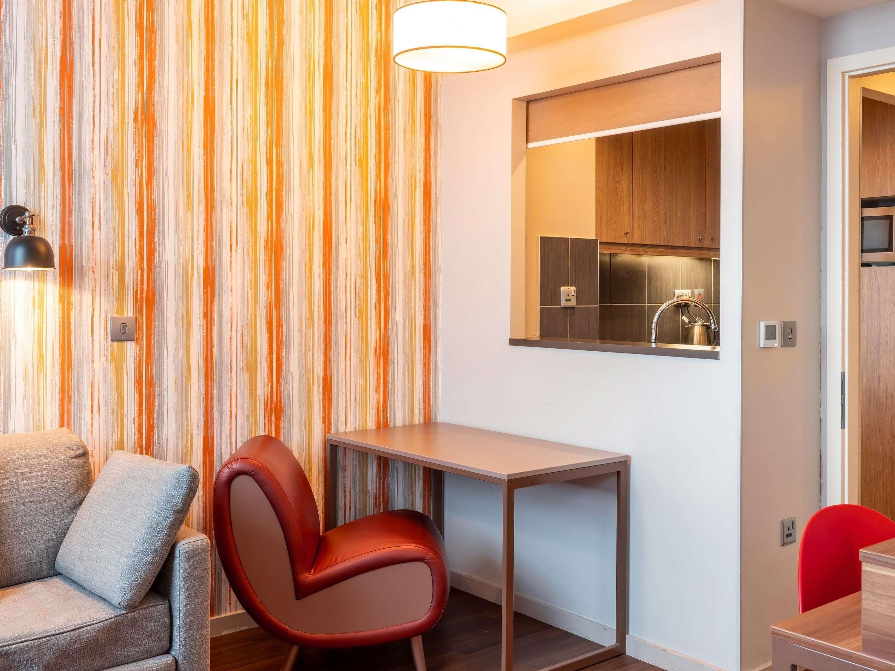 Un espacio habitable compacto con un sofá, una silla roja, una mesita y una kitchenette.
