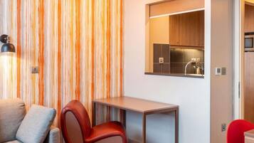 Un espacio habitable compacto con un sofá, una silla roja, una mesita y una kitchenette.