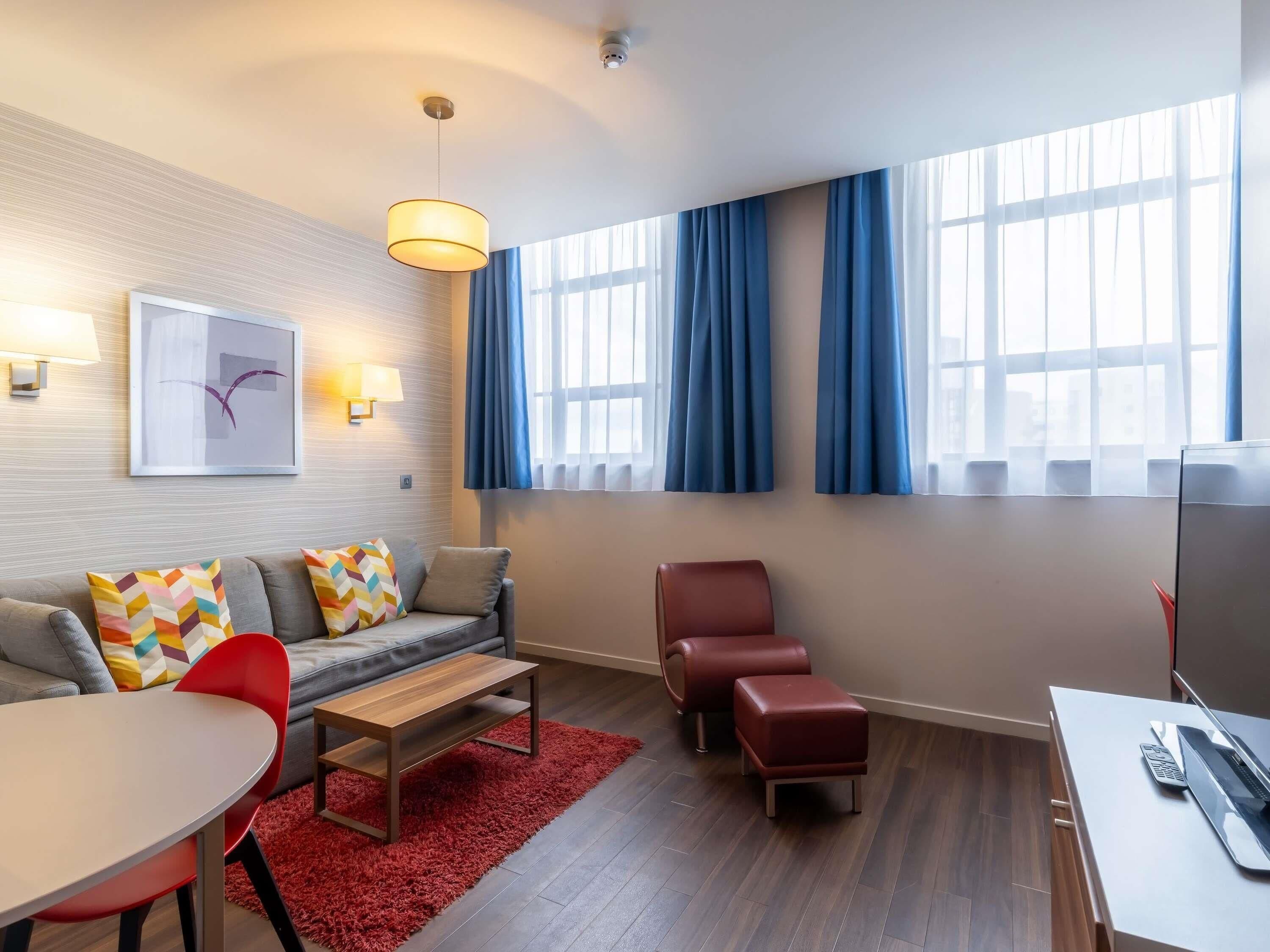 A modern hotel room with a sofa, a red ottoman, a wooden table, a red rug, a desk with a laptop, and a large window with curtains.
