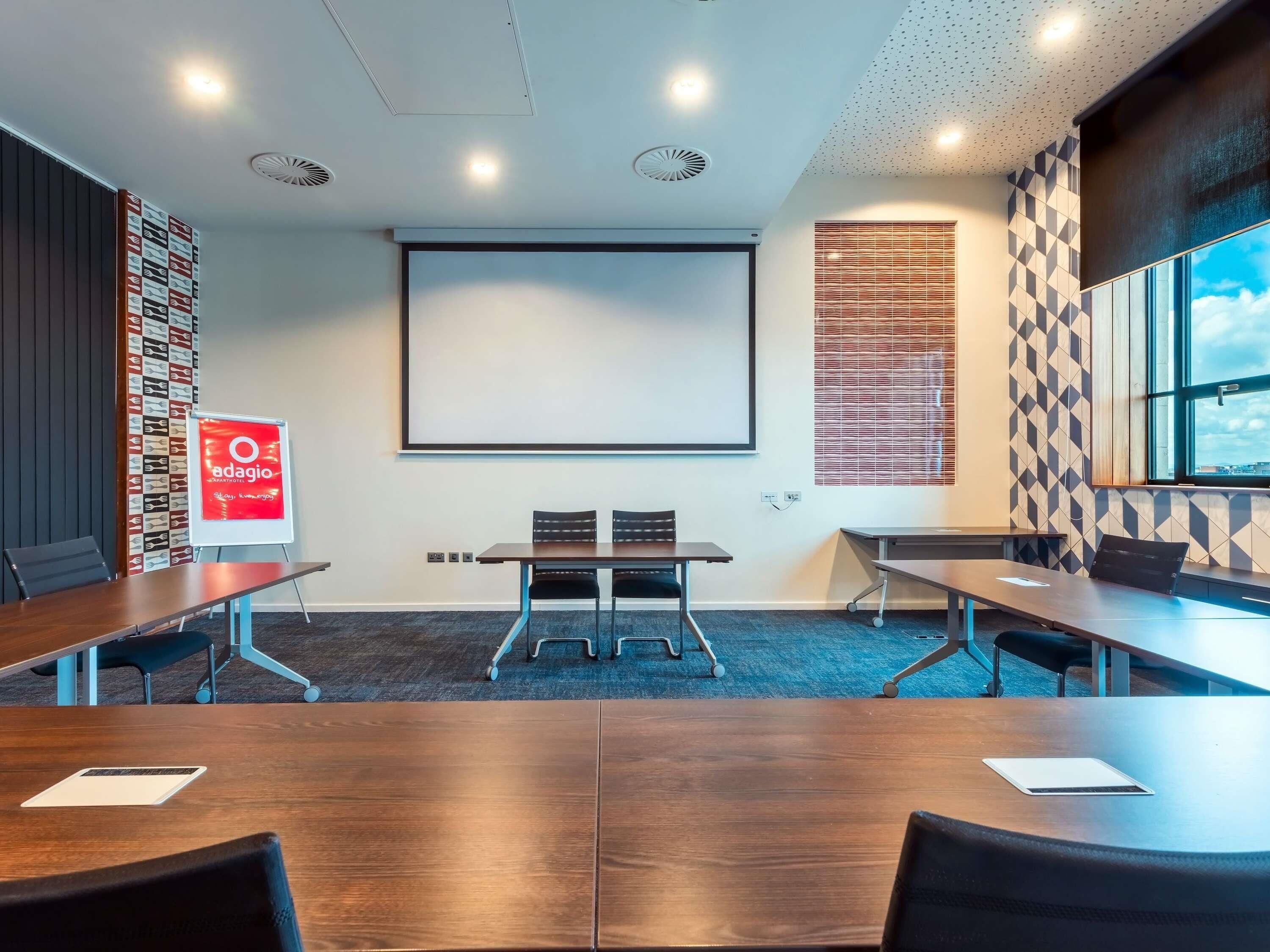 A conference room with a large screen, two tables with chairs, and a patterned wall.