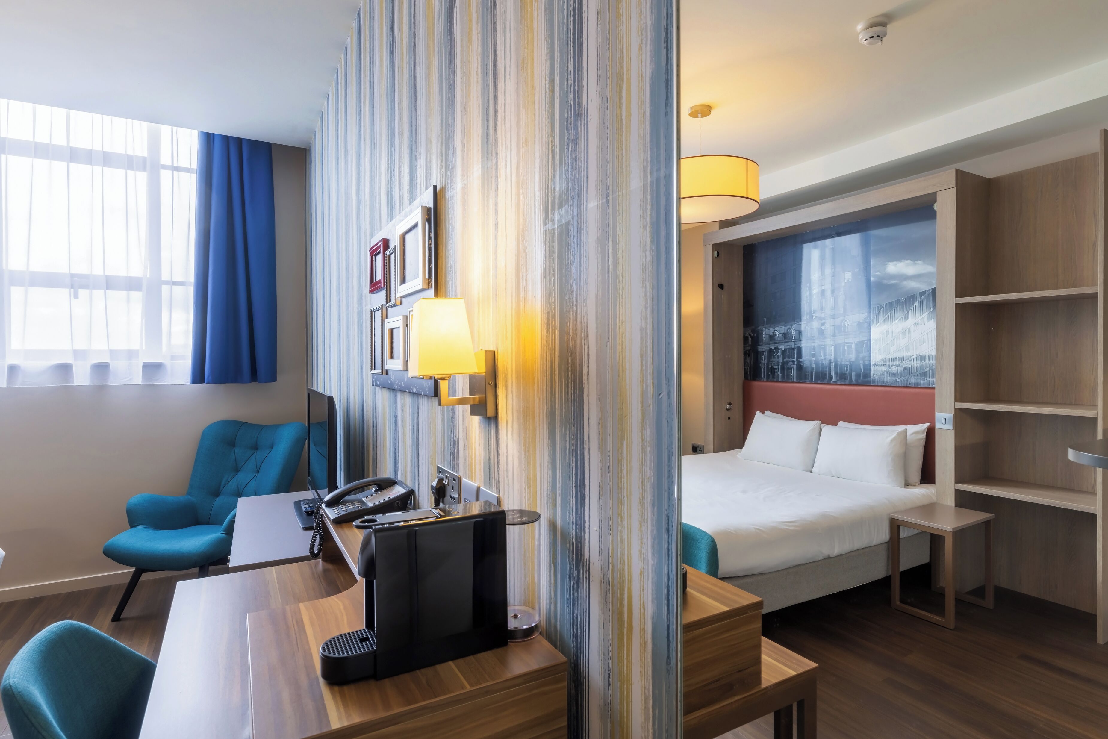A hotel room with a bed, a desk with a computer, a chair, a lamp, and a window with curtains.