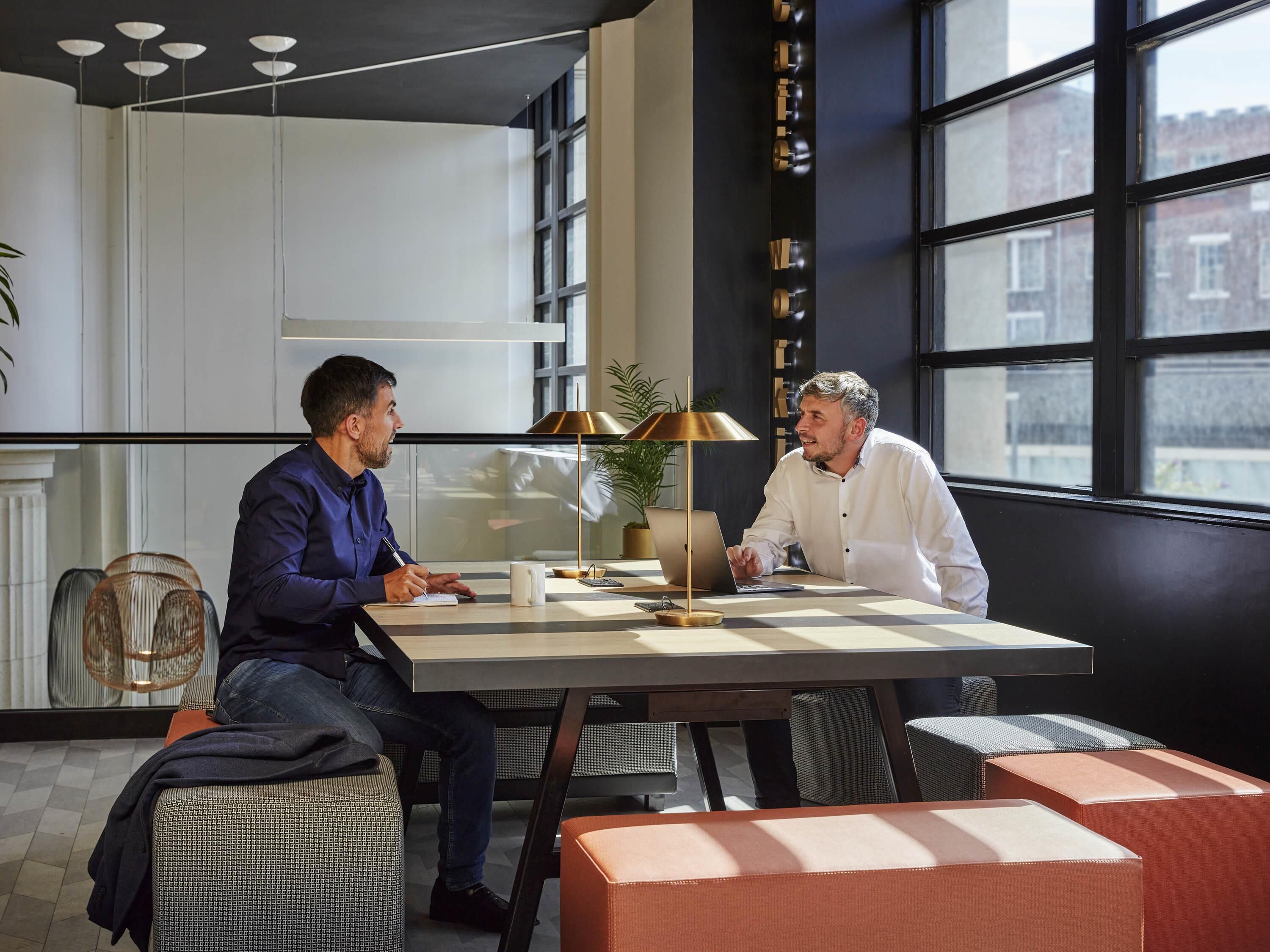 Un bureau moderne avec deux personnes assises à une table, une lampe et une plante en pot.