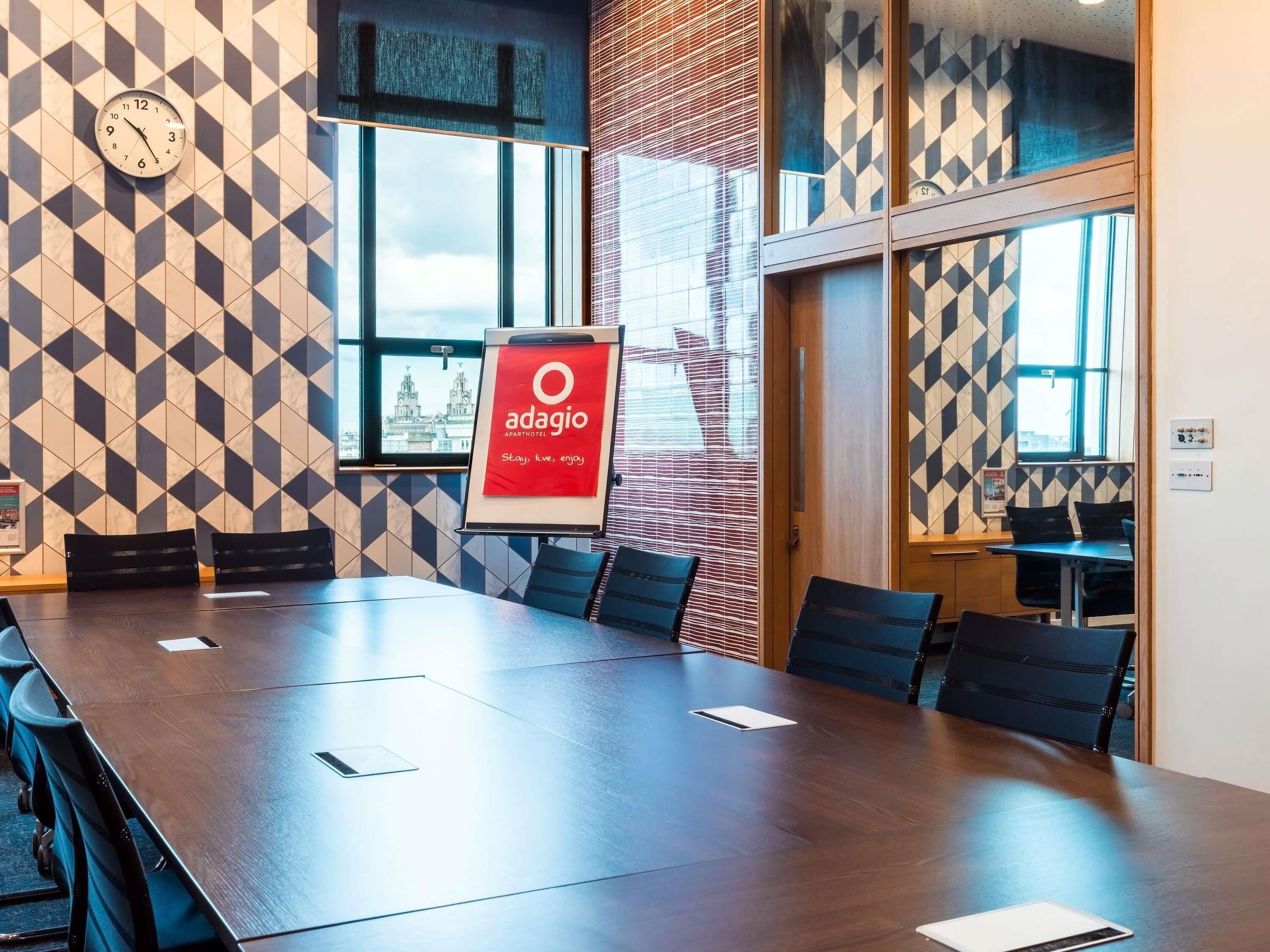 A conference room with a long table, chairs, a clock, and a sign for Adagio apartment hotel.