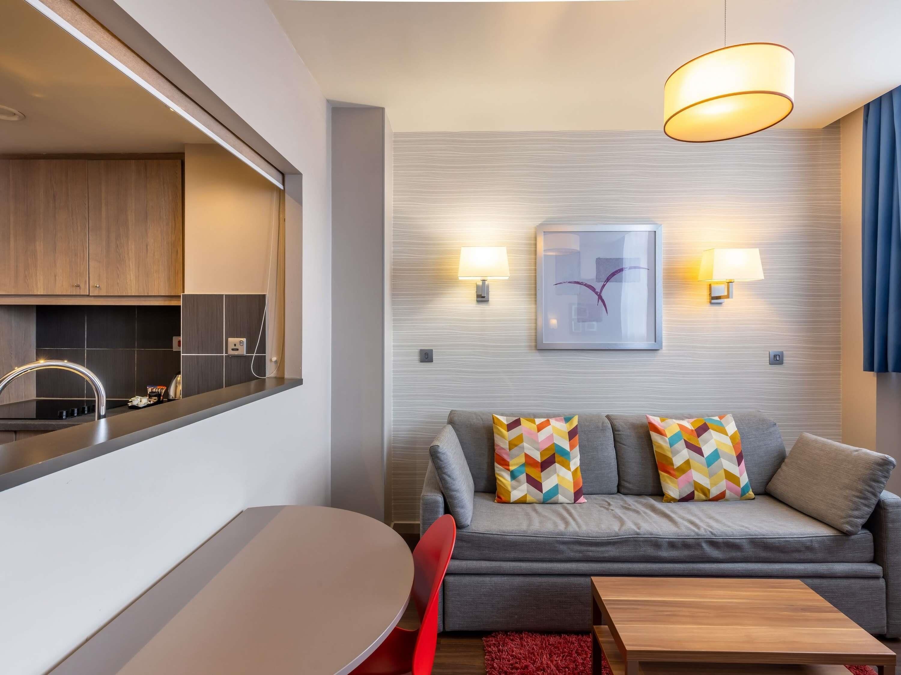 Un salon moderne avec un canapé gris, des coussins colorés, une table basse en bois, un fauteuil rouge et un coin cuisine comprenant un évier et des placards.