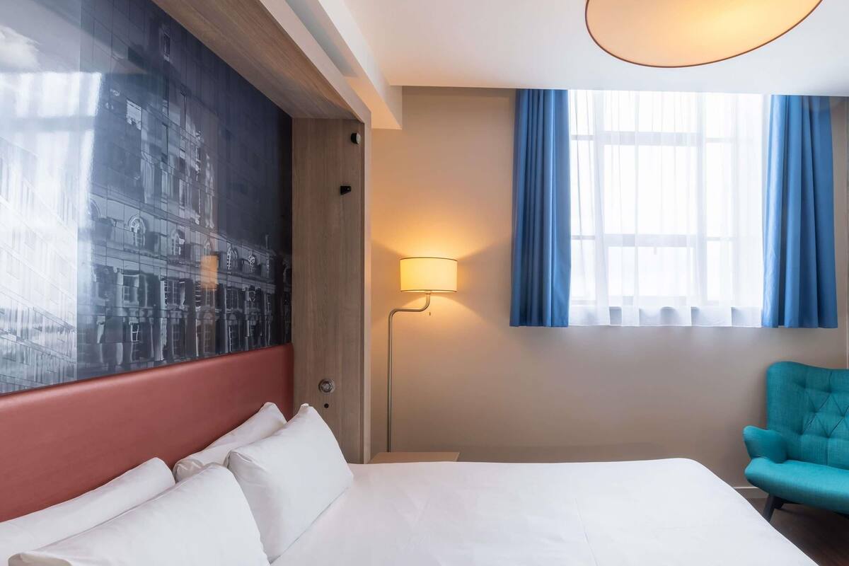 Une chambre d’hôtel avec un grand lit, une chaise bleue, une lampe sur pied et une fenêtre avec des rideaux bleus.