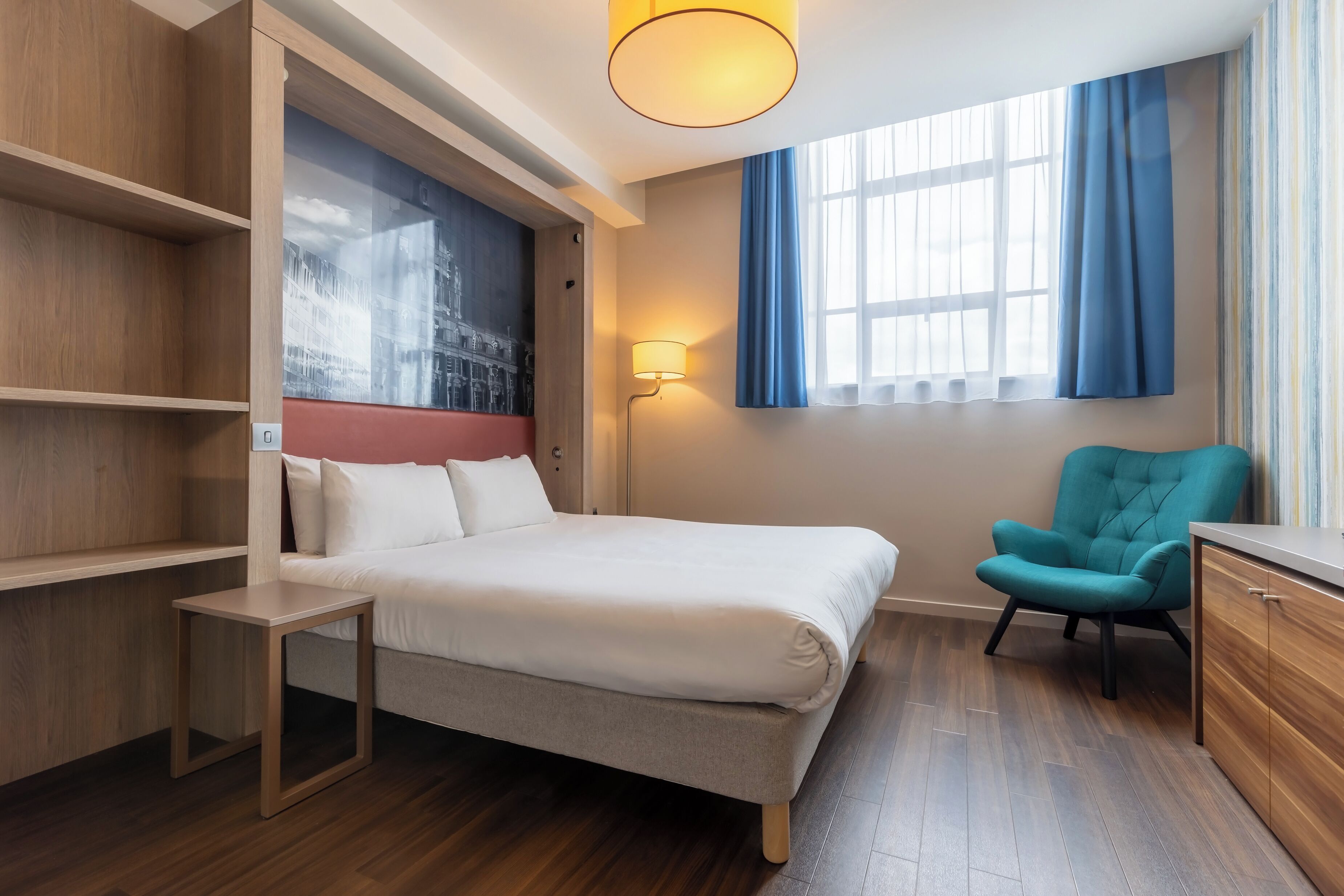A modern hotel room with a bed, a small table, a chair, and a window with curtains.