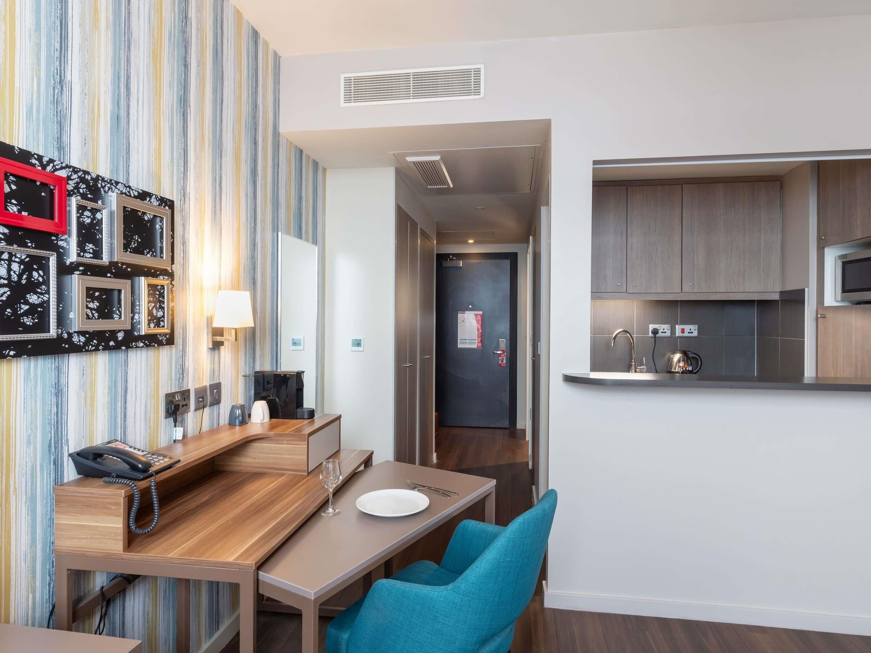 A hotel room with a wooden desk, a telephone, a glass, and a plate. There is a kitchen area with a microwave and a sink.