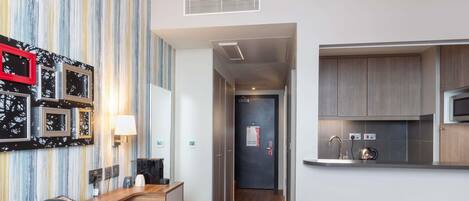 A hotel room with a wooden desk, a telephone, a glass, and a plate. There is a kitchen area with a microwave and a sink.