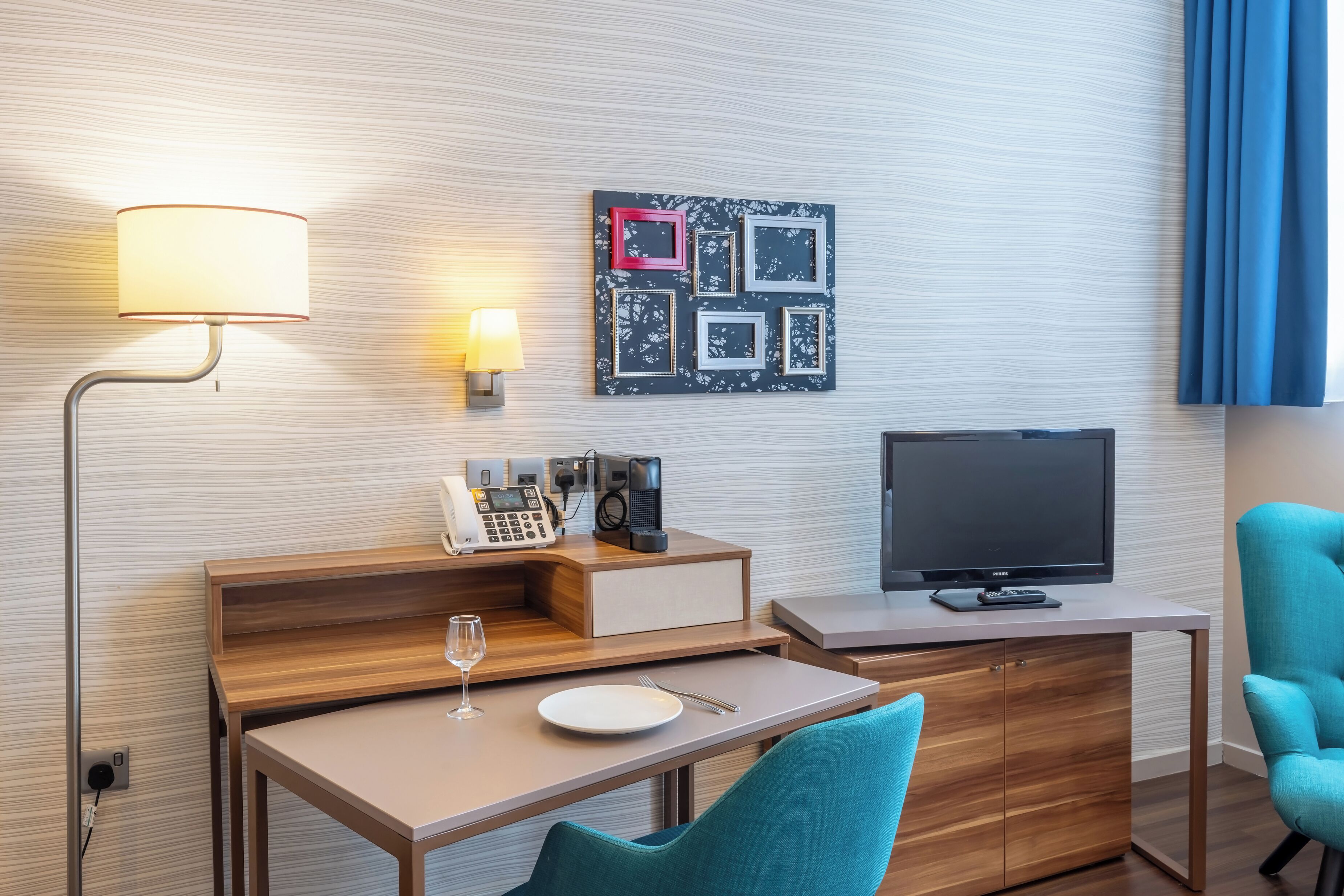 A hotel room with a desk, a chair, a TV, a lamp, a phone, and a wall art piece.