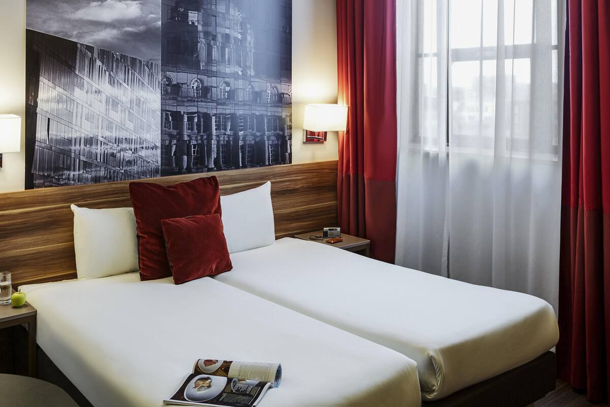 Une chambre d’hôtel avec un grand lit, deux oreillers, une table de chevet, une lampe, un magazine, une fenêtre avec des rideaux et un tableau représentant un paysage urbain accroché au mur.