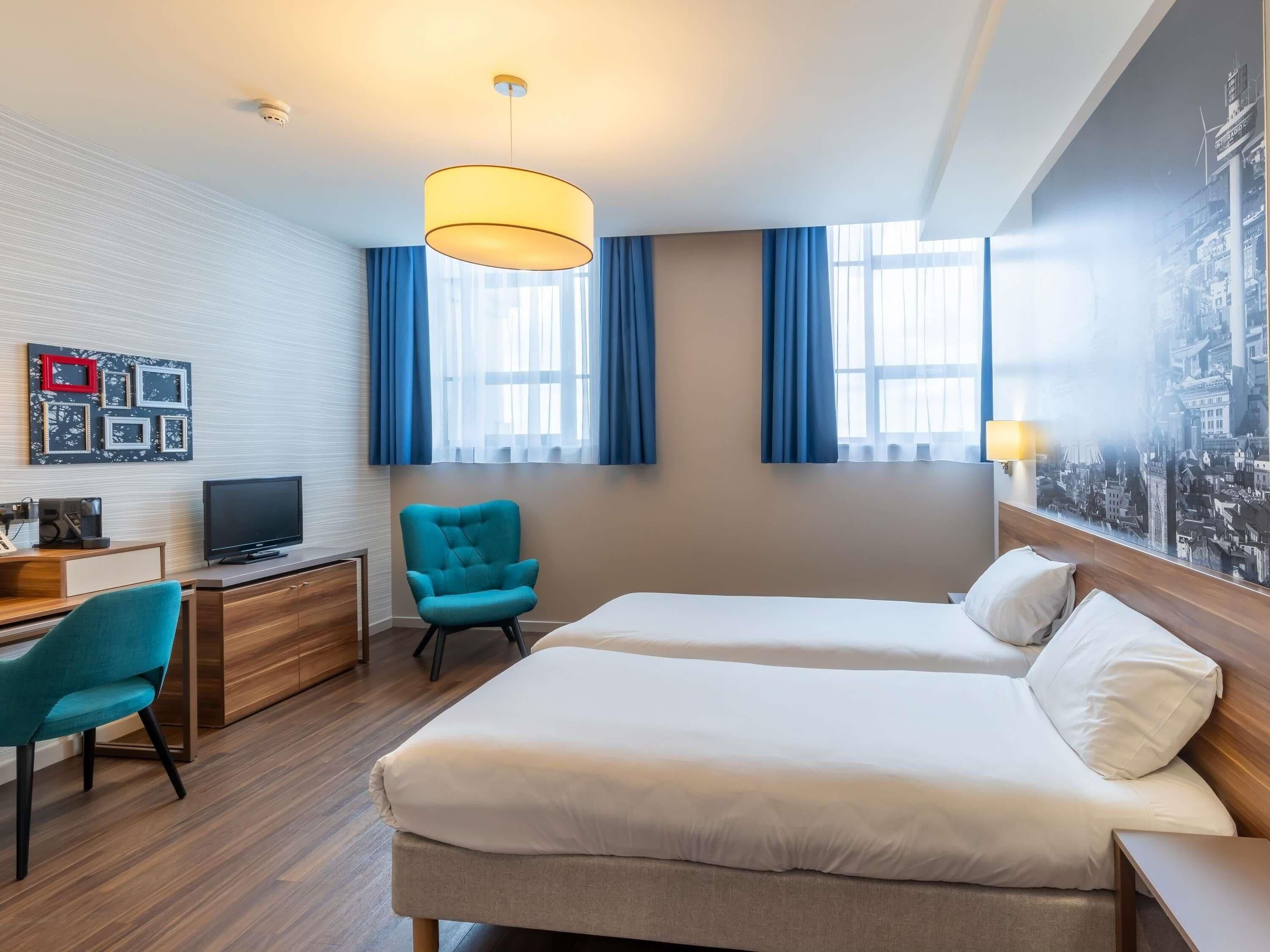 A hotel room with a bed, a desk with a computer, a chair, and a wall-mounted TV.