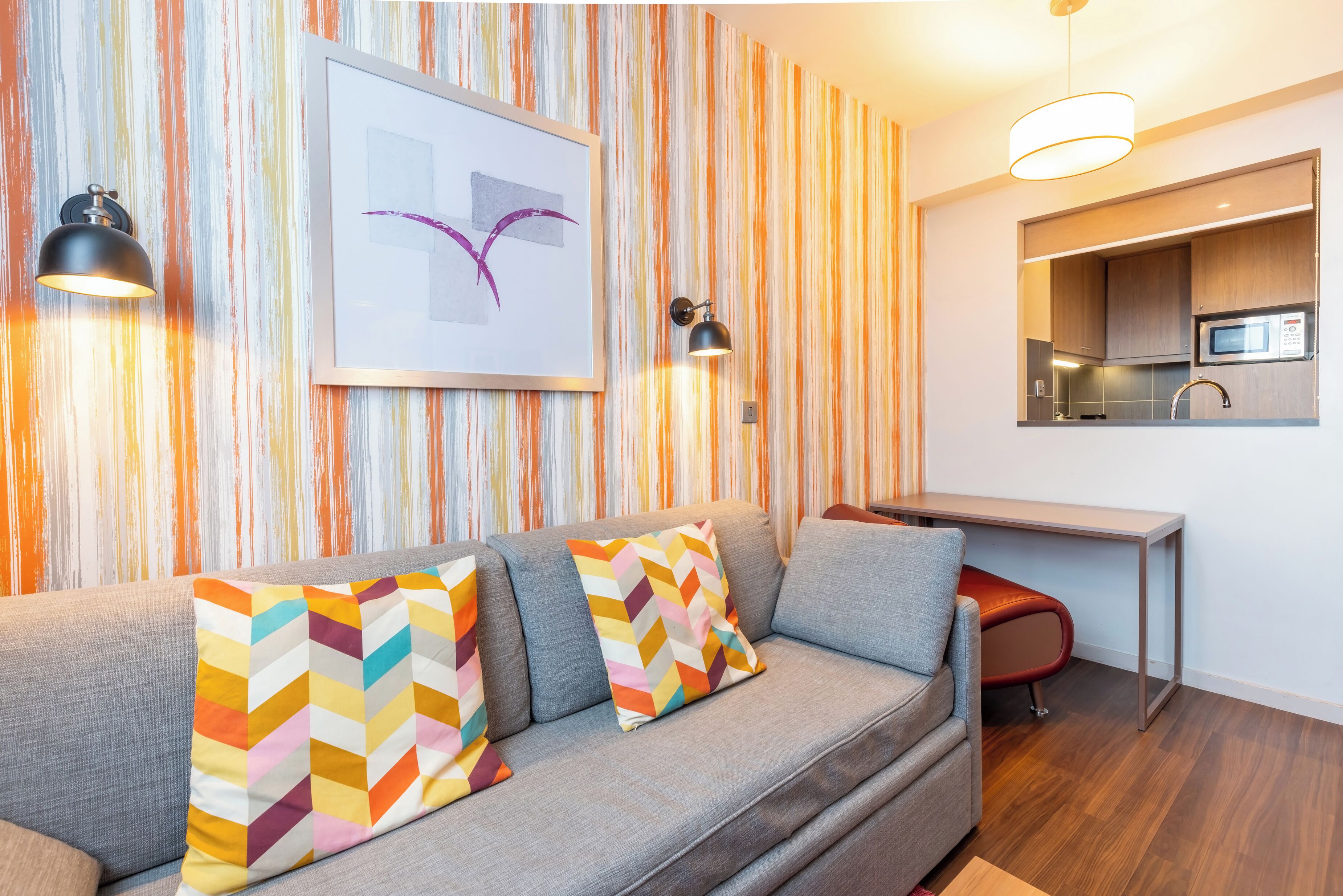 A modern living room with a grey sofa, colorful cushions, a framed artwork, and a kitchen area with a microwave.