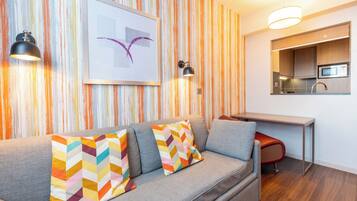 A modern living room with a grey sofa, colorful cushions, a framed artwork, and a kitchen area with a microwave.