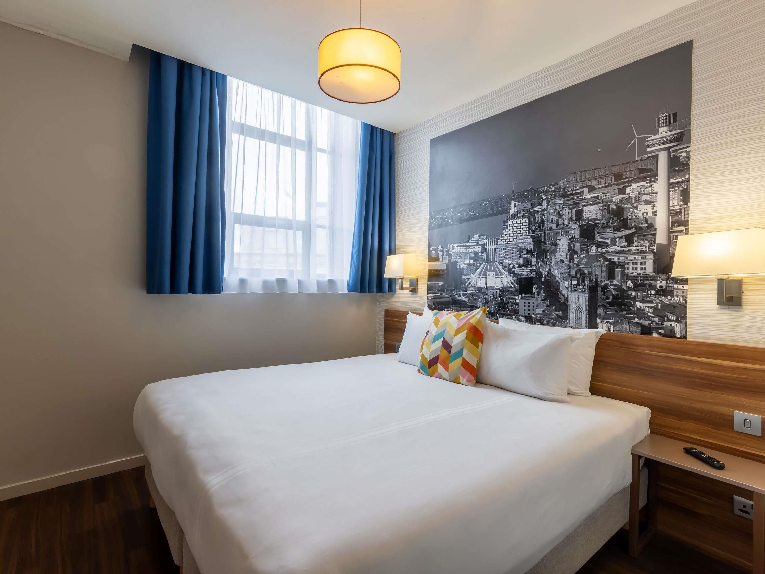 Habitación de hotel con una cama grande, dos lámparas de noche, un ventanal con cortinas azules y un mural con vista a la ciudad.